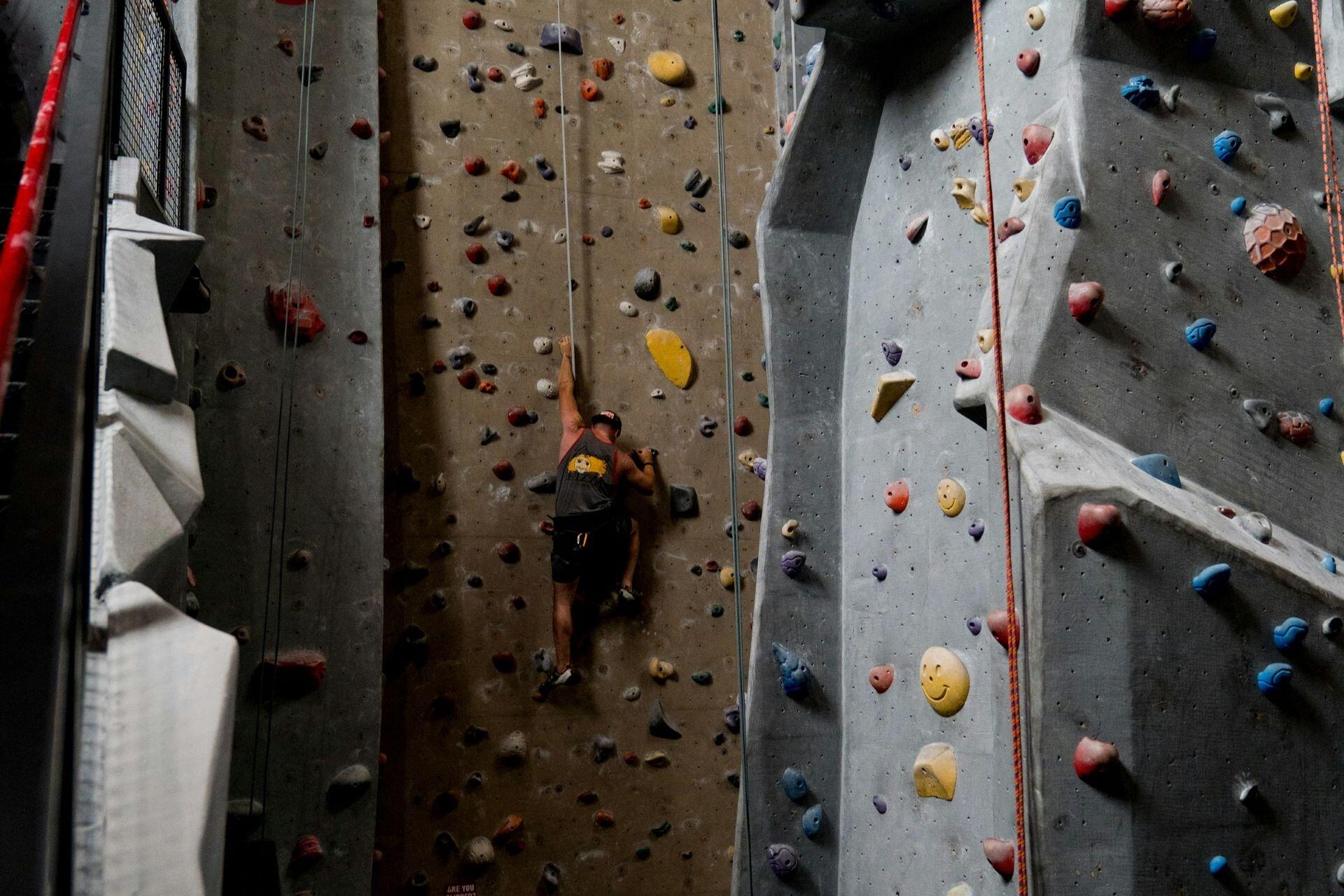 Climber ascending a tall indoor climbing wall surrounded by coloured holds and ropes
