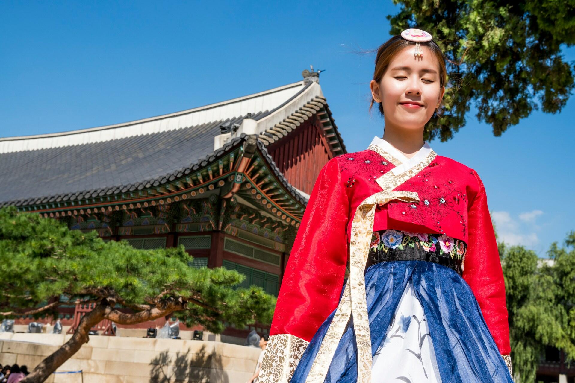 A young girl with black hair pulled back wears a red hanbok top with a blue skirt on a sunny day.