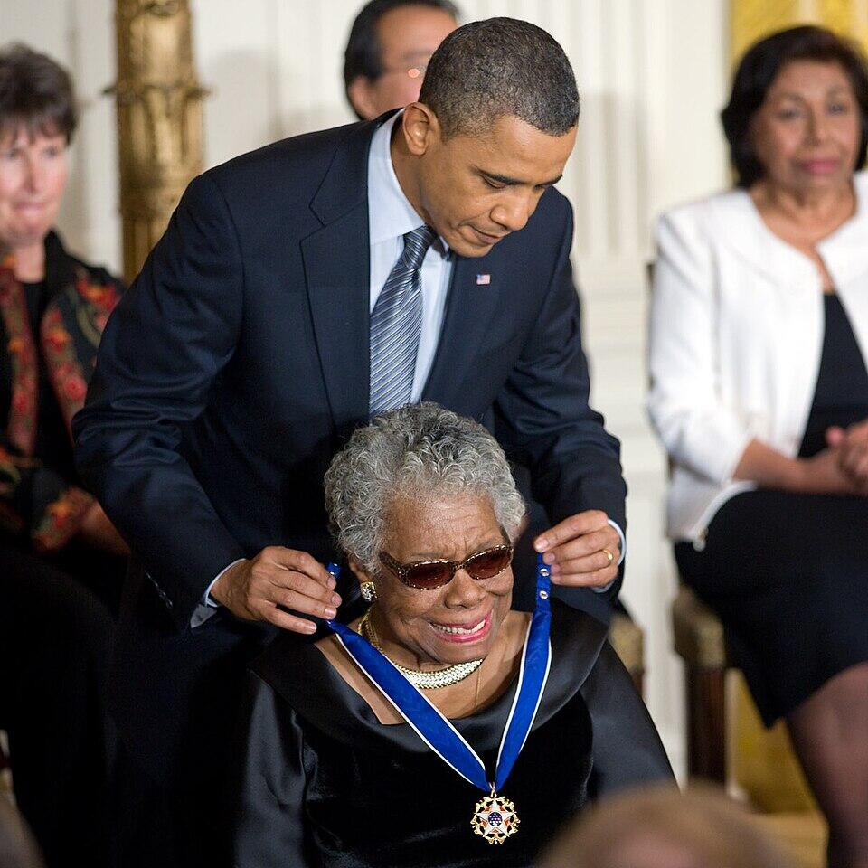 Barack Obama places a blue-ribboned medal around Maya Angelou’s neck as seated guests watch.