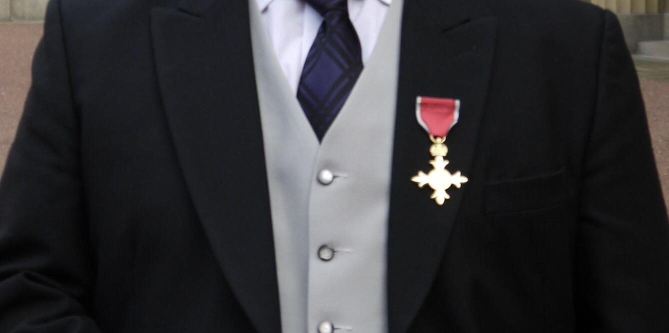 A medal shaped in a golden cross, hanging by a short red ribbon trimmed in gold, adorns the lapel of a suit jacket.