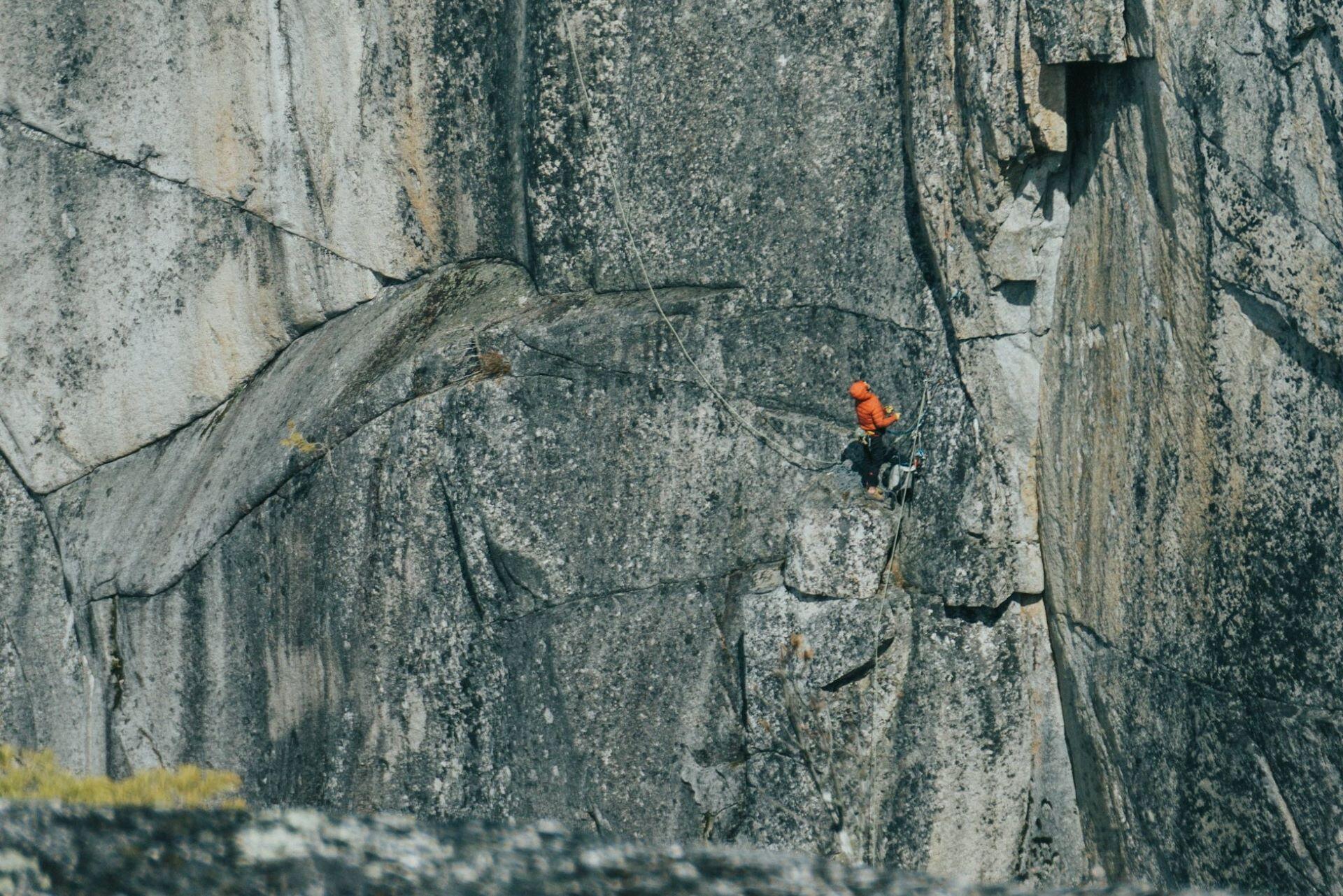Climber in an orange jacket ascending a large outdoor rock face with ropes