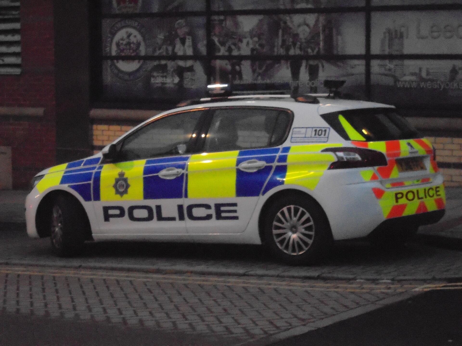 A white car with yellow and blue decals, with the word 'police' stencilled in blue, sits parked at the kerb on a city street.