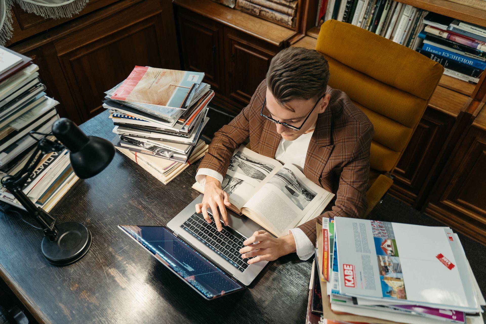 A man in glasses and a brown plaid jacket types on a laptop at a desk surrounded by stacks of books and papers.