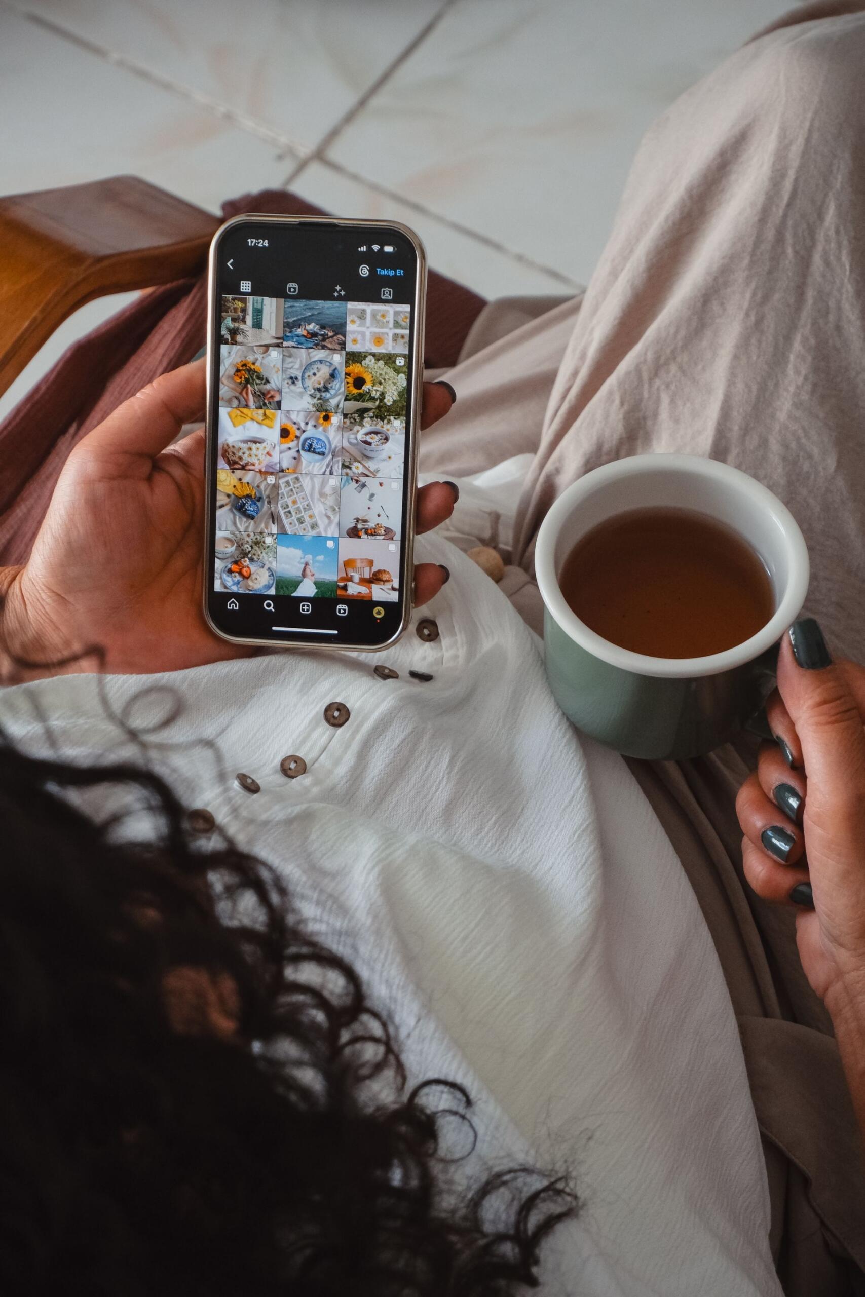A seated person in a white shirt holds a phone showing a grid of photos and a green mug, with dark nail polish.