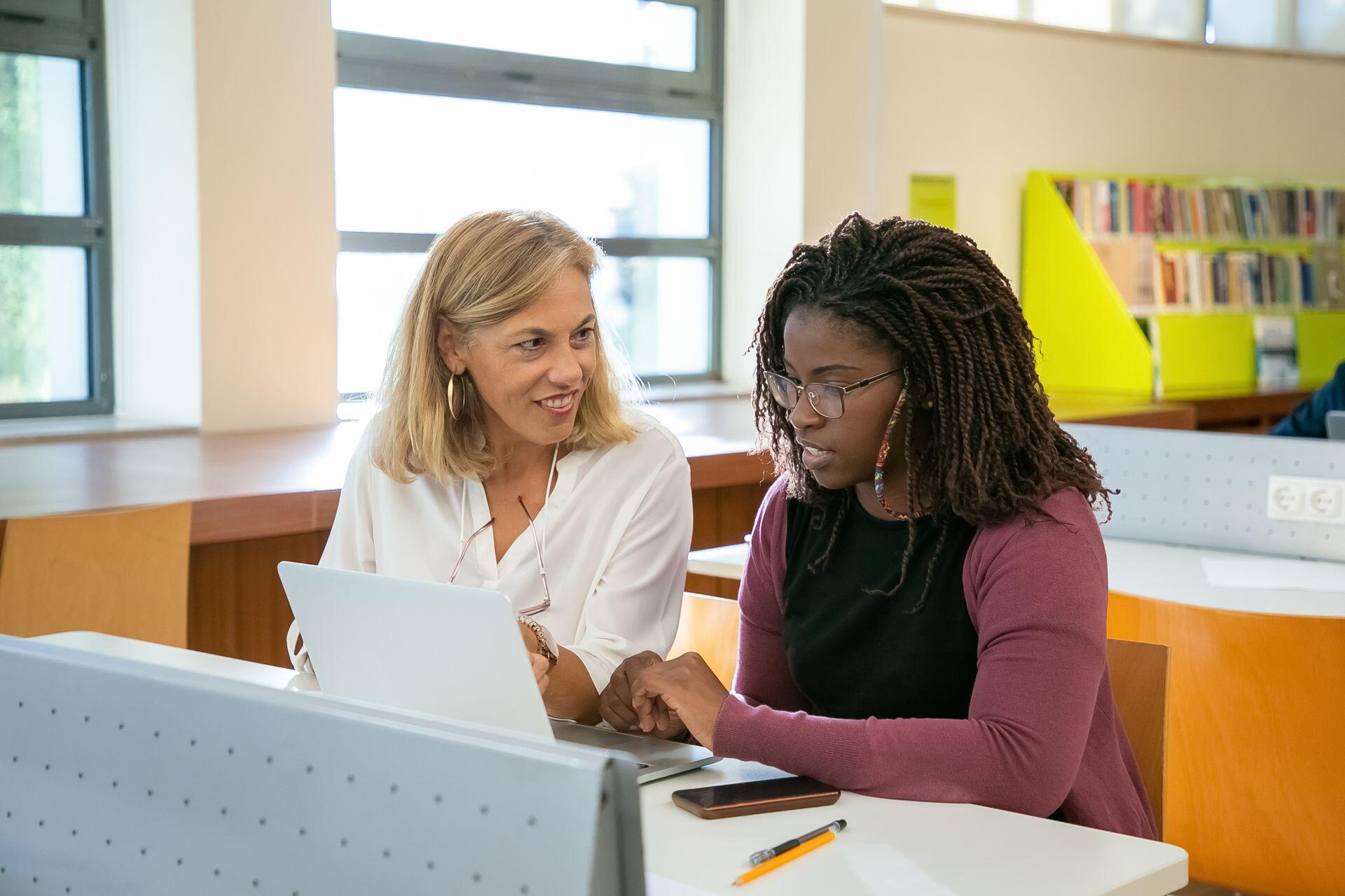 Two women sit at a table in a library, looking at a laptop beside a phone and pencil, with bookshelves behind them.