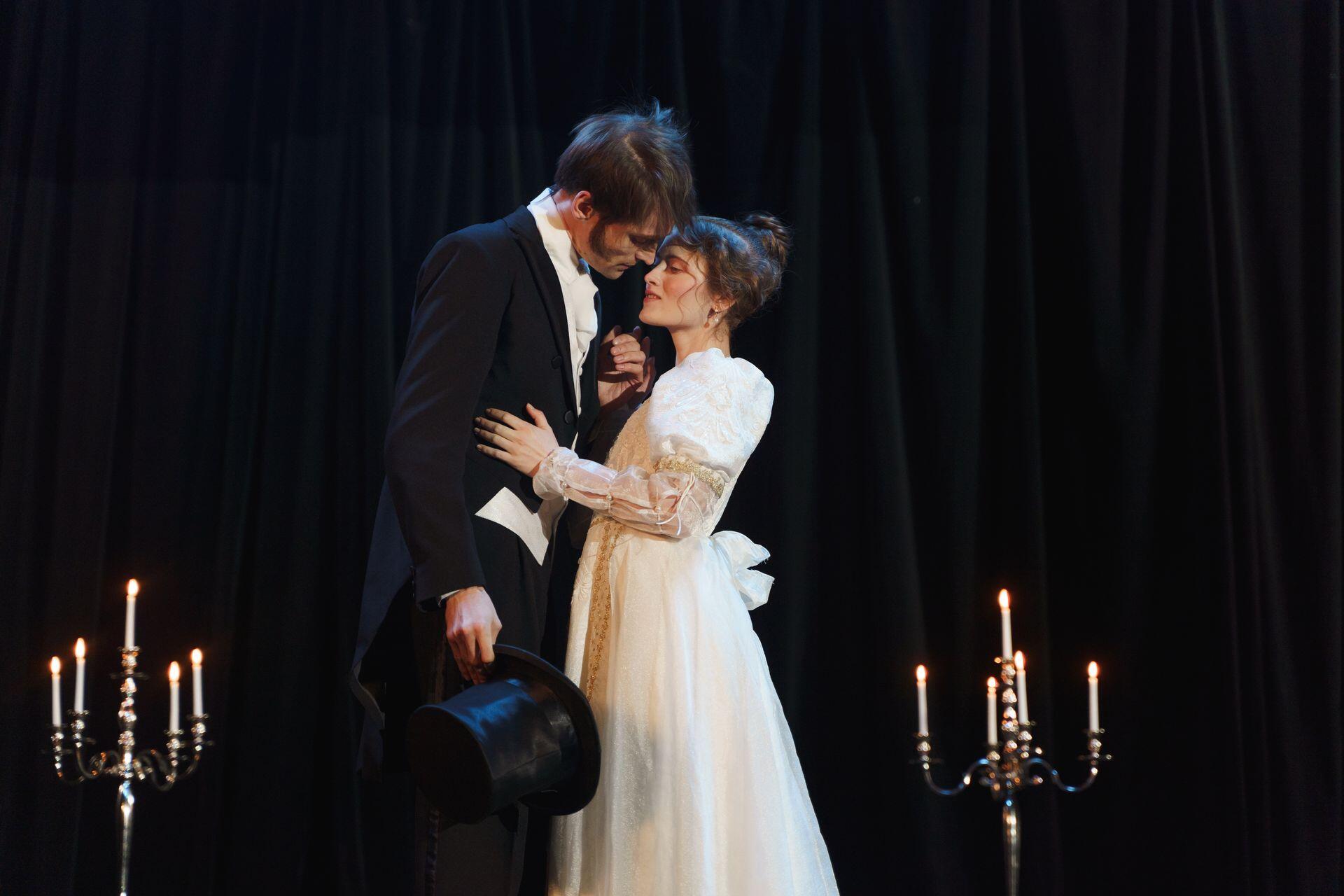 A man in a black suit and a woman in a white dress stand close together on a stage, surrounded by candlelit candelabras.