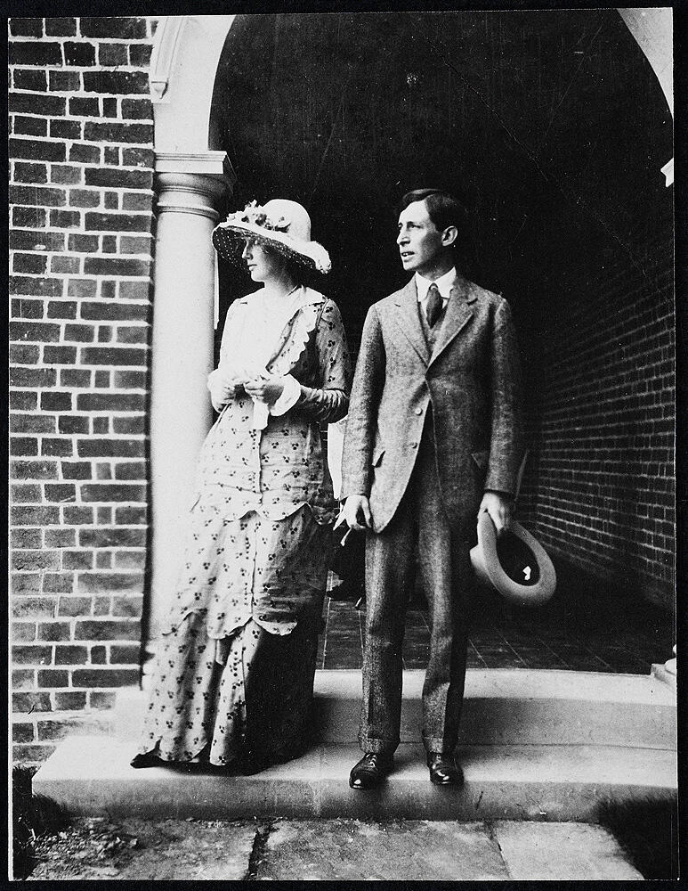 A man in a suit holding a hat and a woman in a long dress and wide-brimmed hat stand beneath a brick archway.
