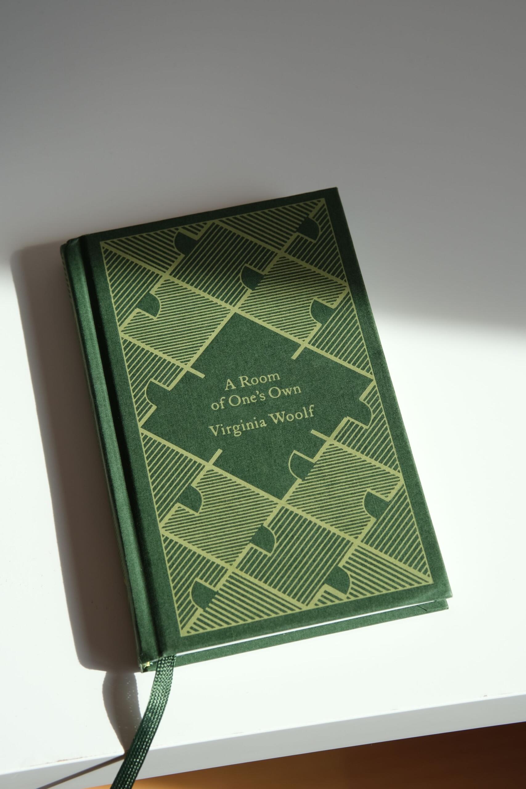A green hardcover book titled “A Room of One’s Own” by Virginia Woolf lies diagonally on a white surface with a ribbon bookmark.