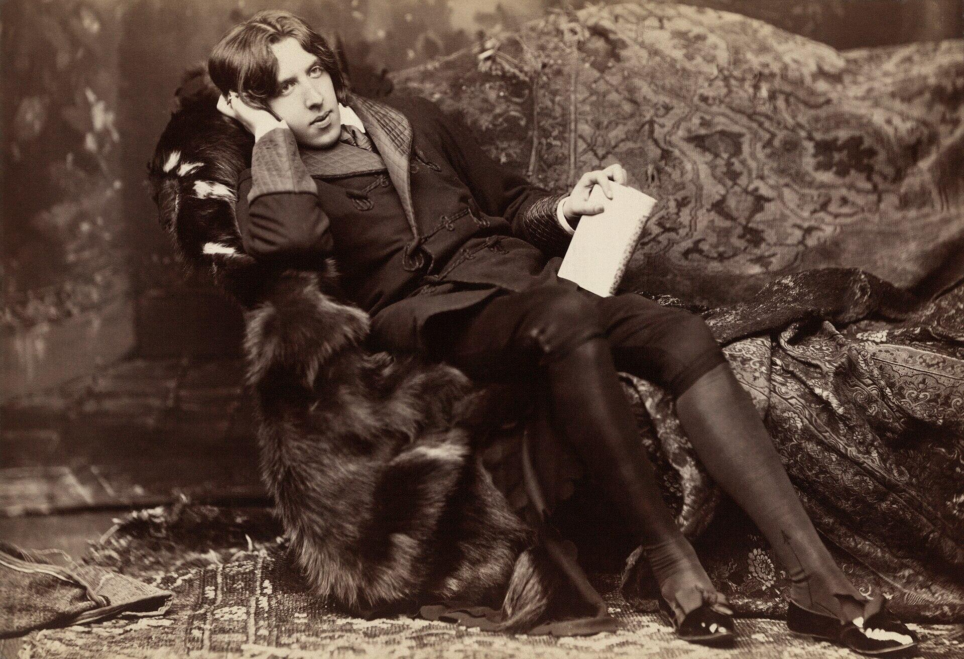 A sepia portrait shows a young man reclining on a patterned sofa, wearing a dark suit and holding a sheet of paper.