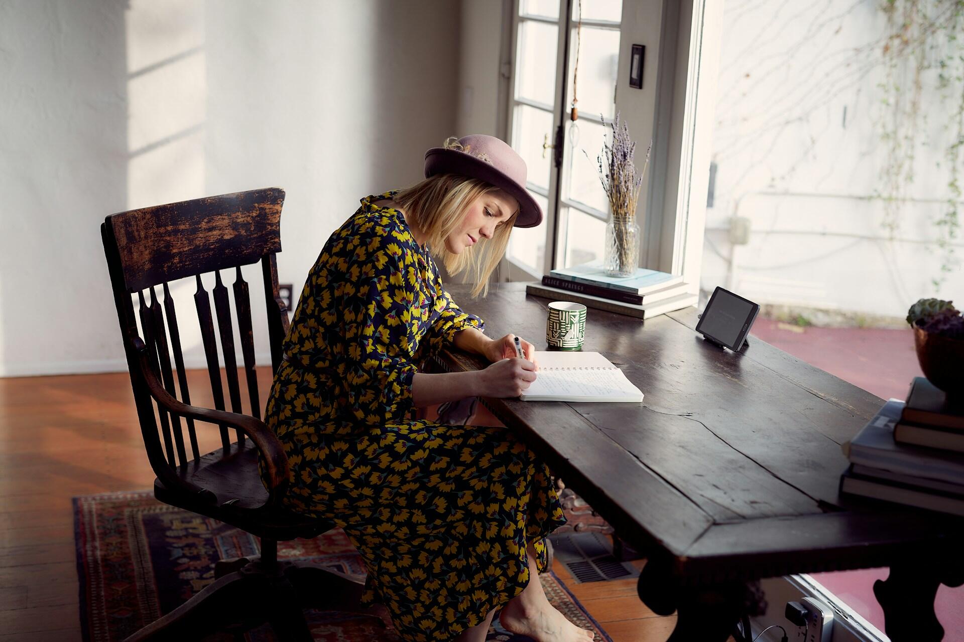 A woman in a floral dress and pink hat writes in a notebook at a wooden table beside a sunlit window.