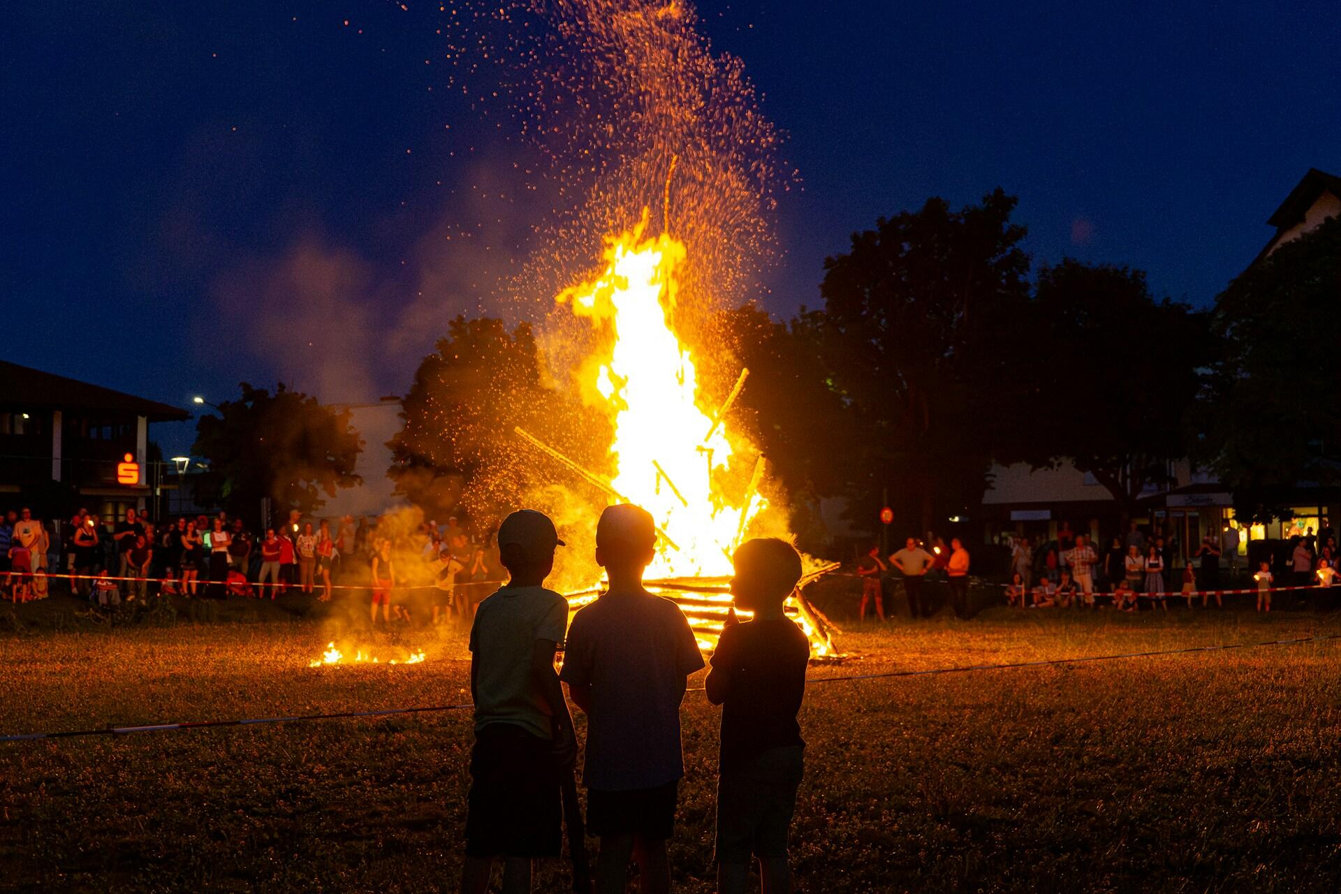 Three children stand in front of a bonfire while a crowd of people stand in the background. The mood is festive and solemn.