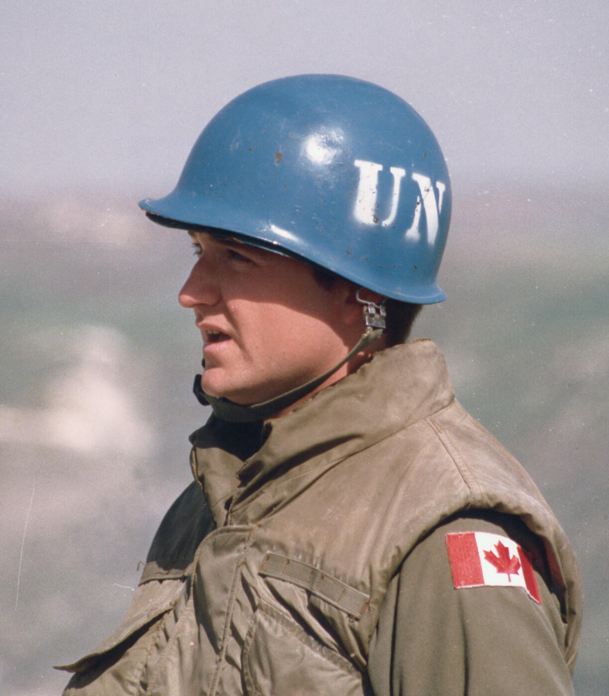 A man in a tan uniform and vest wears a blue helmet marked “UN” and a Canadian flag patch on his sleeve.