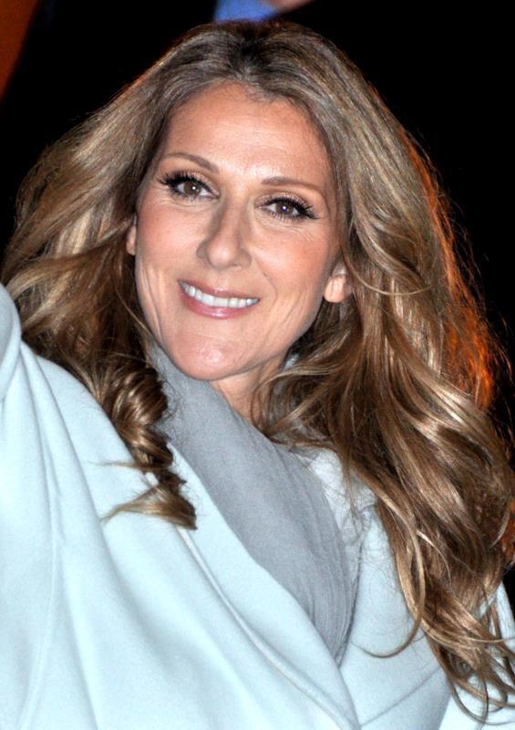 A smiling woman with long wavy light-brown hair wears a pale blue coat and gray scarf against a dark background.