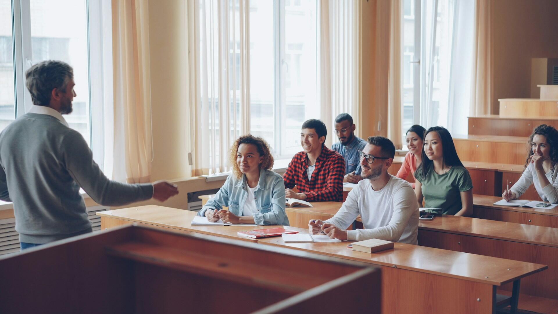 A man in a grey jumper addresses a small group of students in a classroom. The mood is friendly but studious.