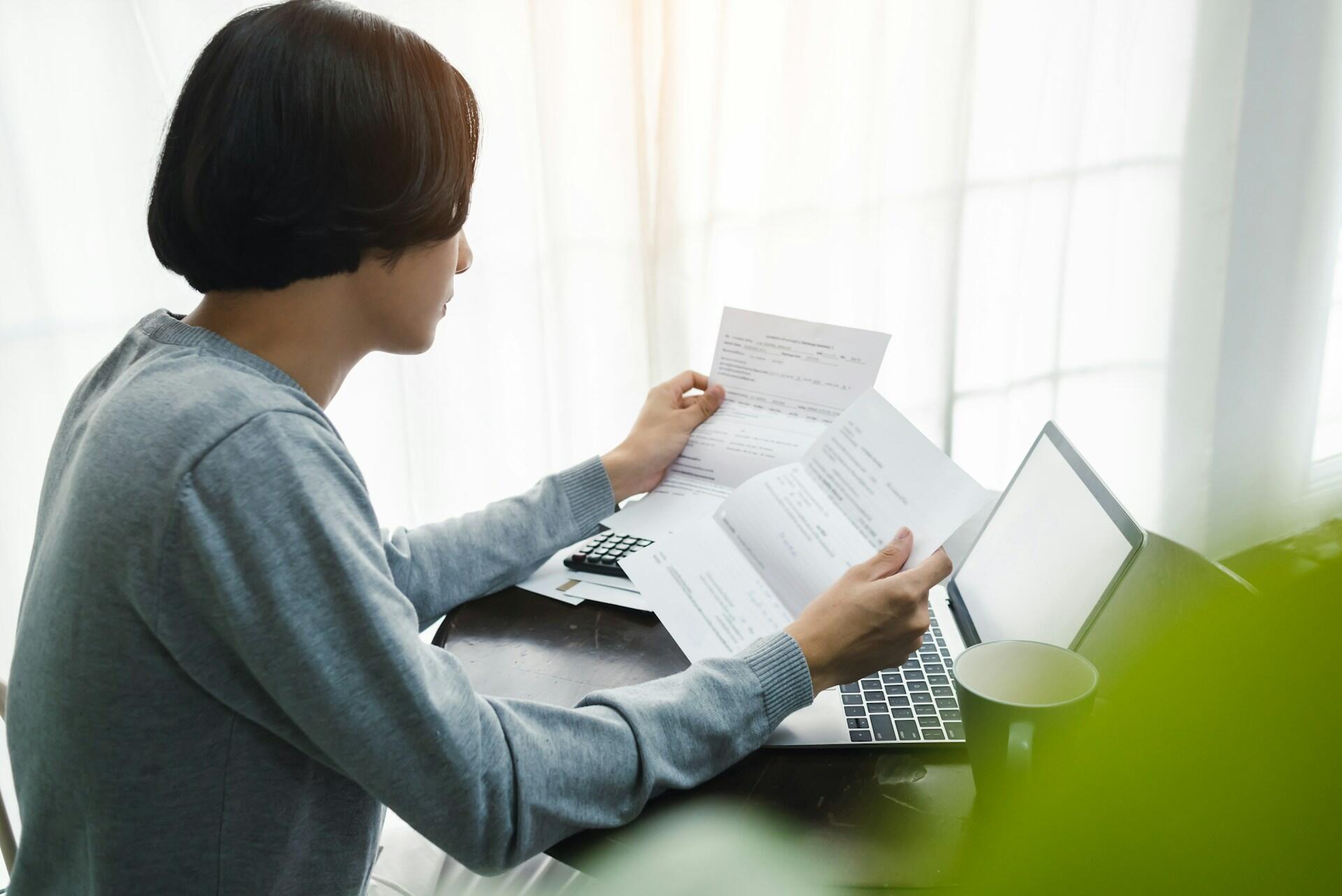 A young woman sits at a table holding a sheet of paper in each hand,, focused on the left-hand page. The mood is anxious and worried.