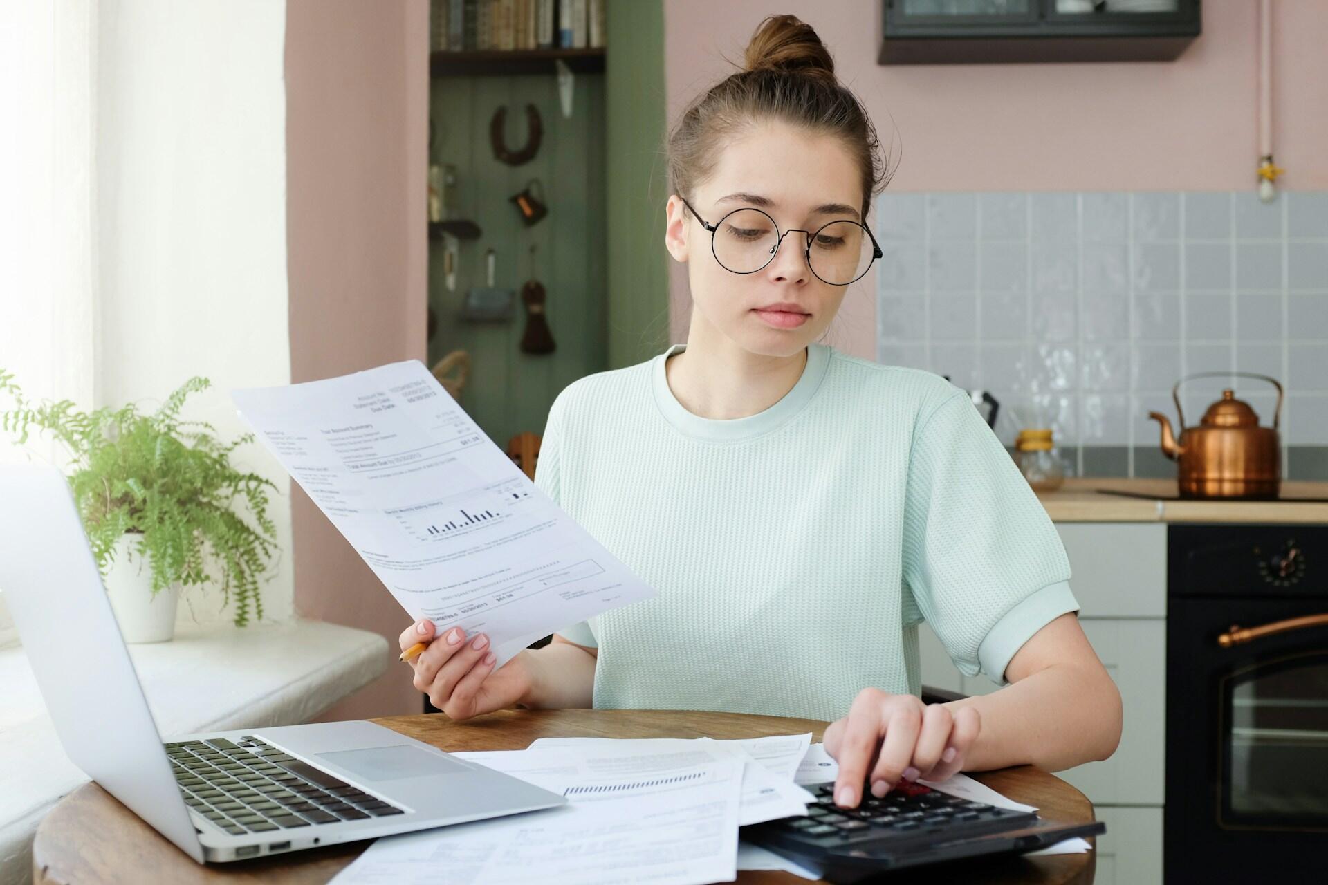 A young woman wearing glasses holds a sheet of paper up while working a calculator. The mood is studious and thoughtful.