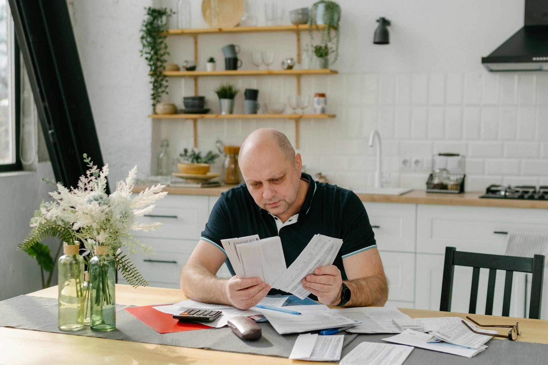 A man wearing a black shirt sits at a table laden with paperwork, holding three sheets of paper. The mood is serious and concerned.