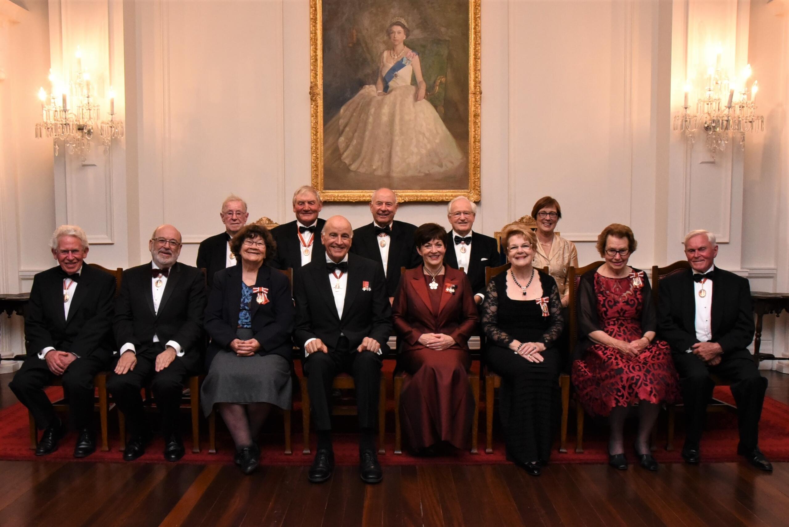 A group of thirteen older adults in formal evening clothes pose in two rows beneath a large portrait in an ornate room.