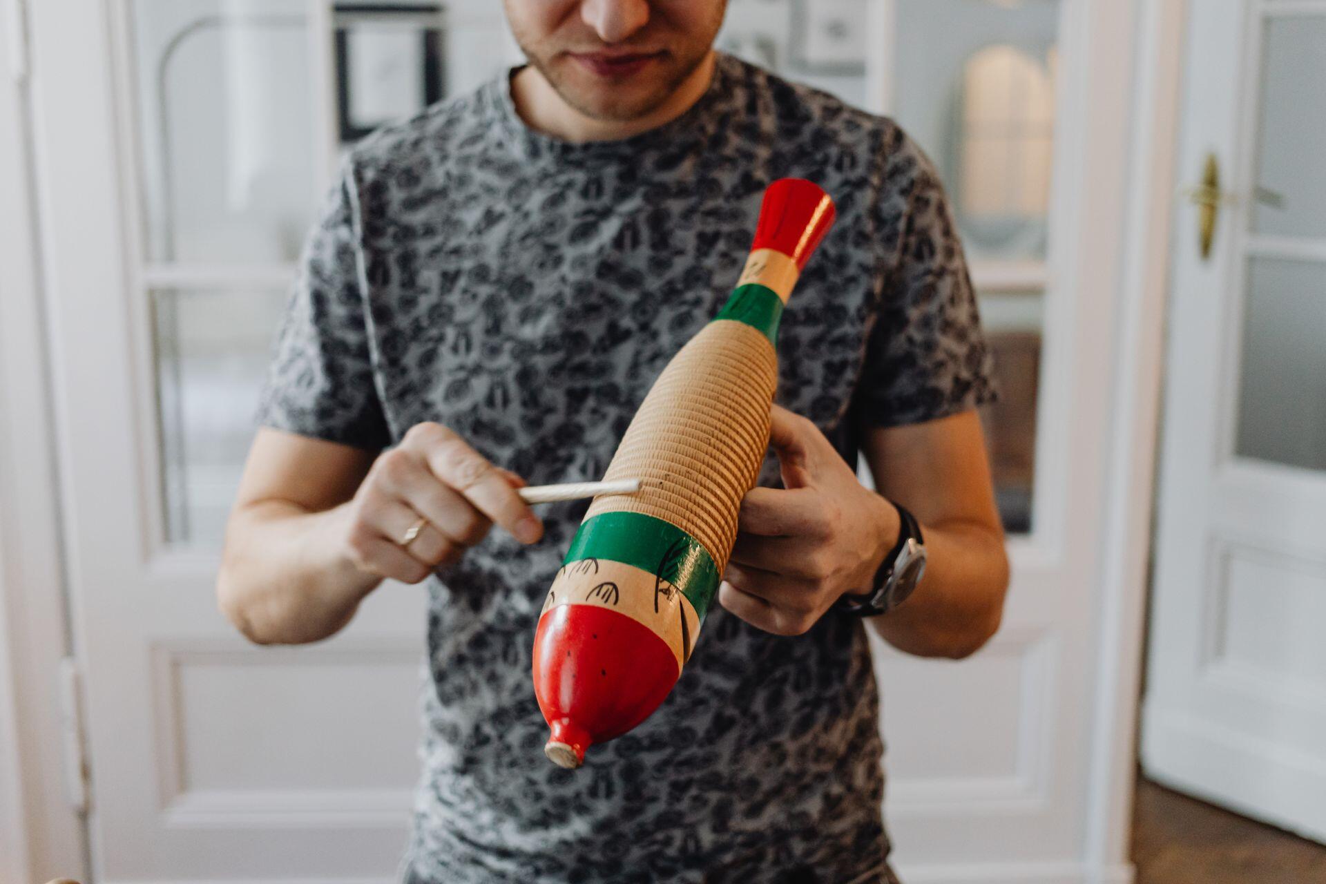 person holding a wooden guiro with red and green painted stripes on each end, running a wooden stick along the ridges