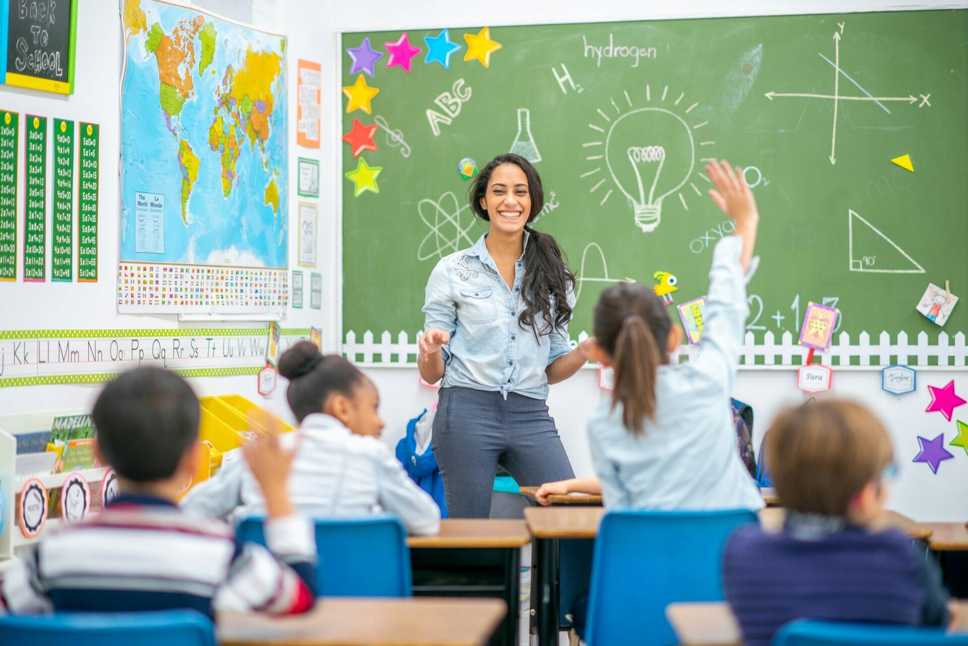 A woman with long hair stands in a classroom with children seated at their desks, one with their hand up. The mood is happy and excited.