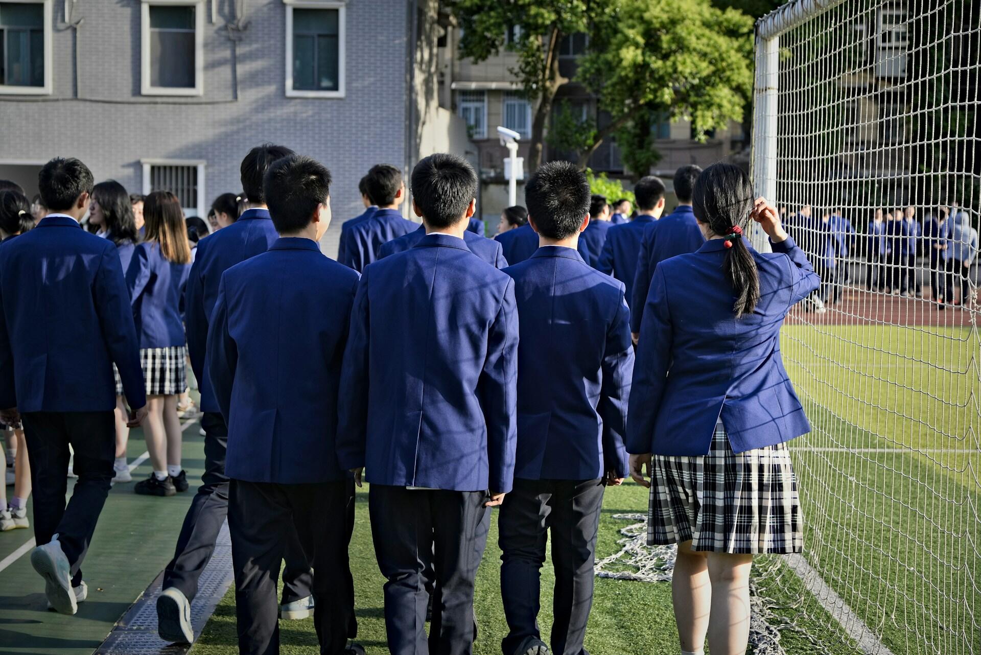 Students in blue school uniforms walk along a sports field beside a tall net fence, with a school building and trees behind them.