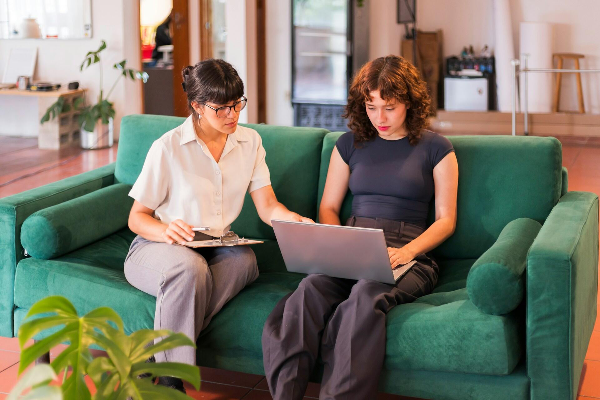 Two women sit on a sofa, one with an open computer on her lap and the other pointing to the screen. The mood is serious and studious.