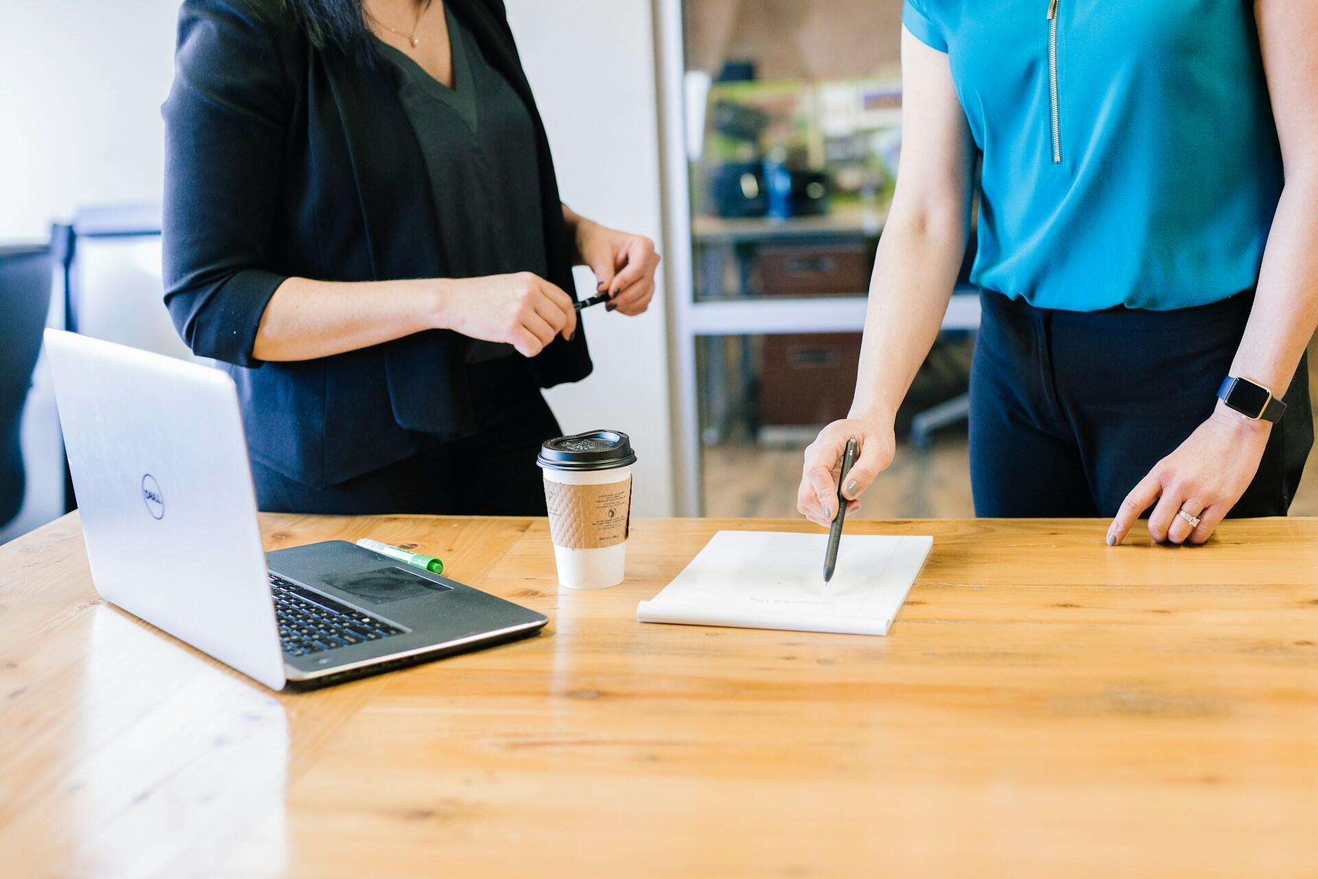Two people standing by a table in an office, each with a pen in hand. The mood is constructive and collaborative.