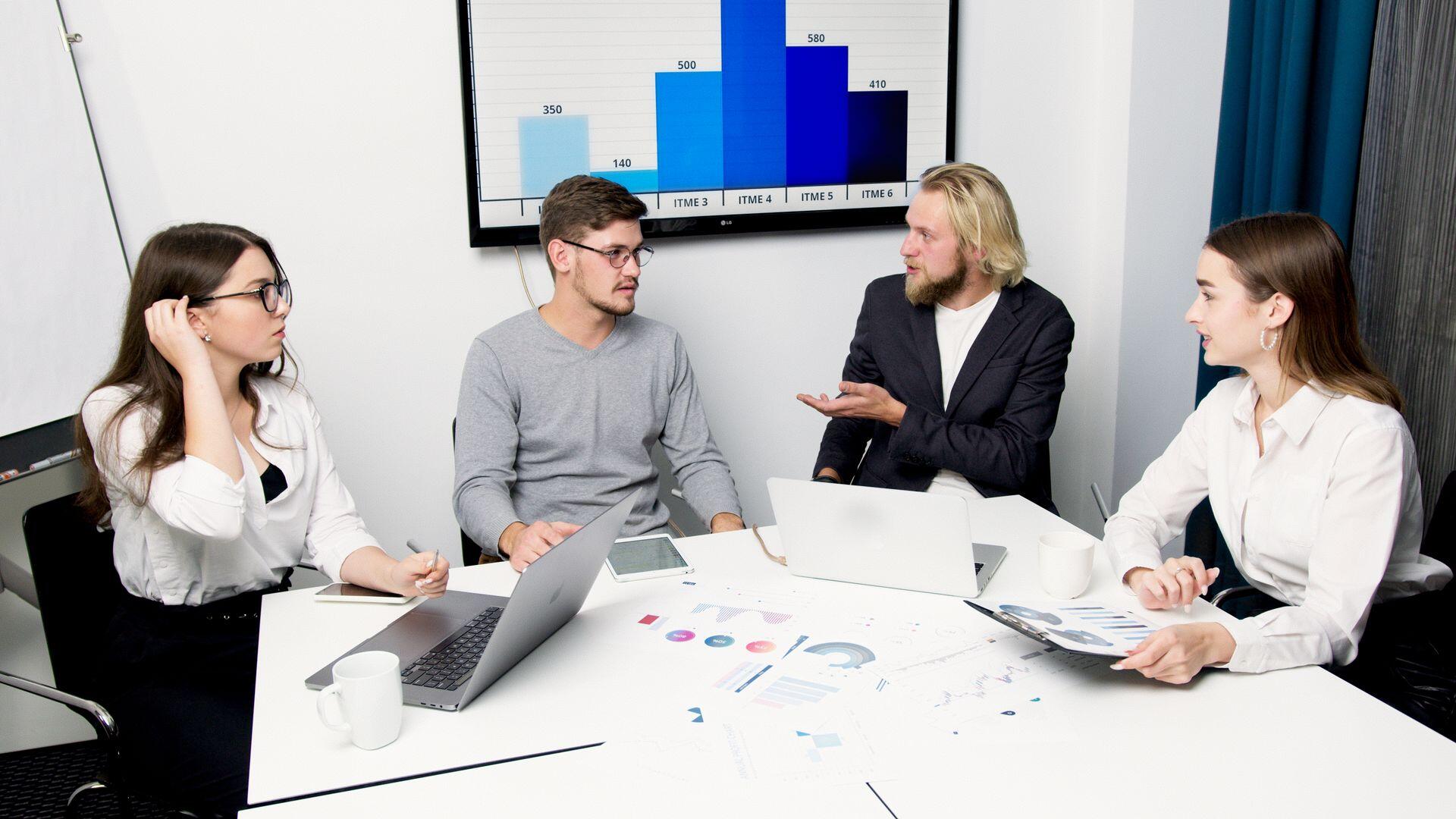 Four coworkers discuss charts around a conference table with laptops and printed reports, beneath a screen showing a blue bar graph.