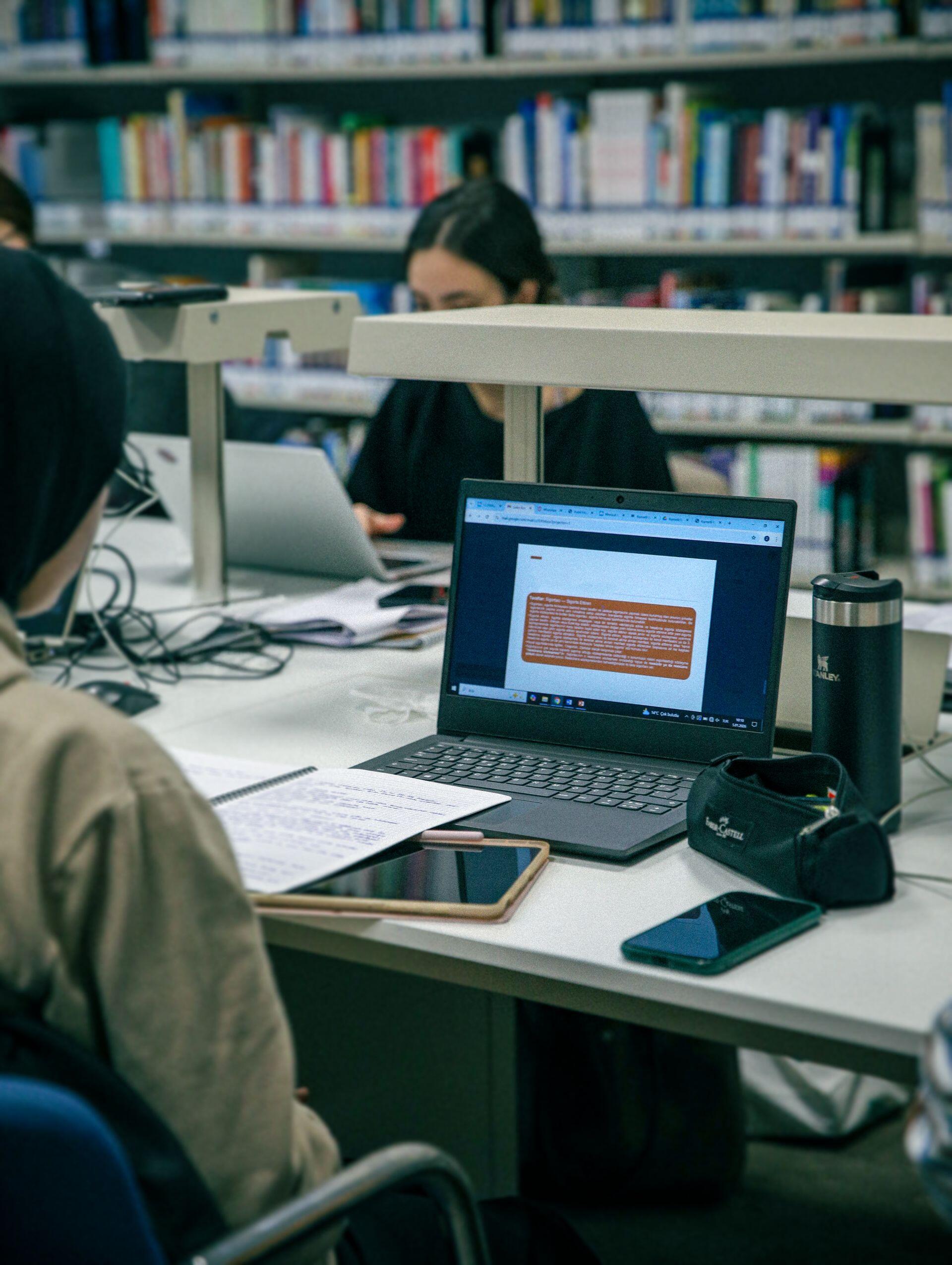 Students study at a library table with an open laptop, papers, phones, a pencil case, and a thermos, with bookshelves behind them.