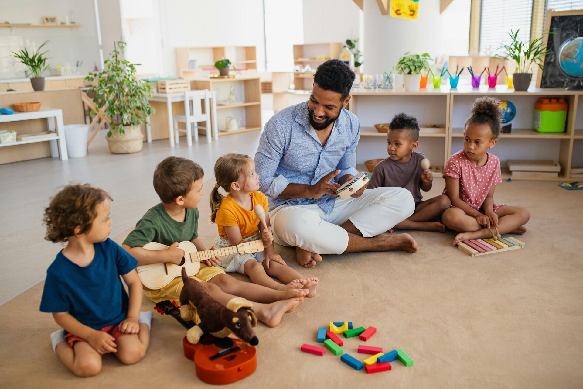 A bearded man sits on the floor, surrounded by small children, holding a tambourine. The mood is happy and excited.