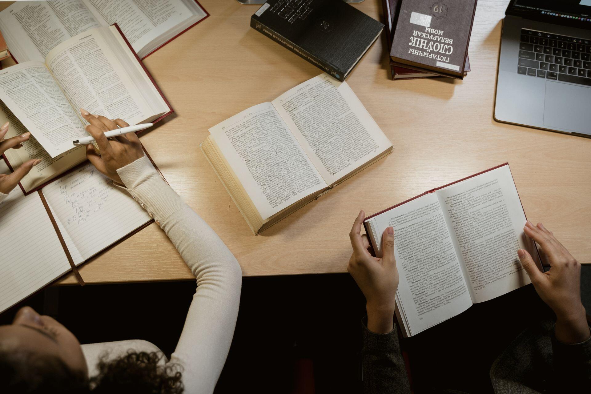 Two people read open books at a wooden table, with a notebook, stacked books, and part of a laptop nearby.