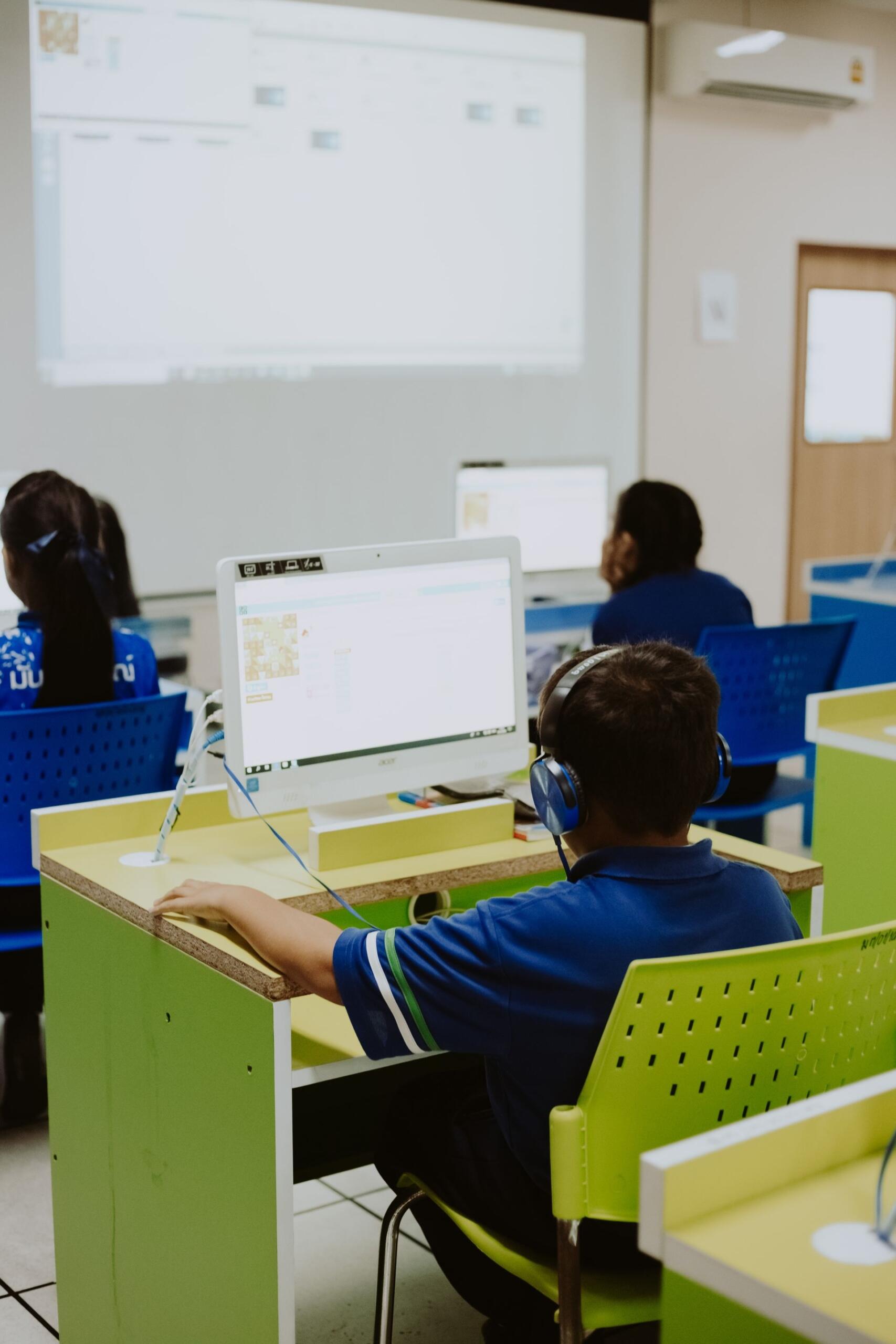 A student in a blue uniform and headphones sits at a computer in a classroom lab, with other students and a projected screen behind him.