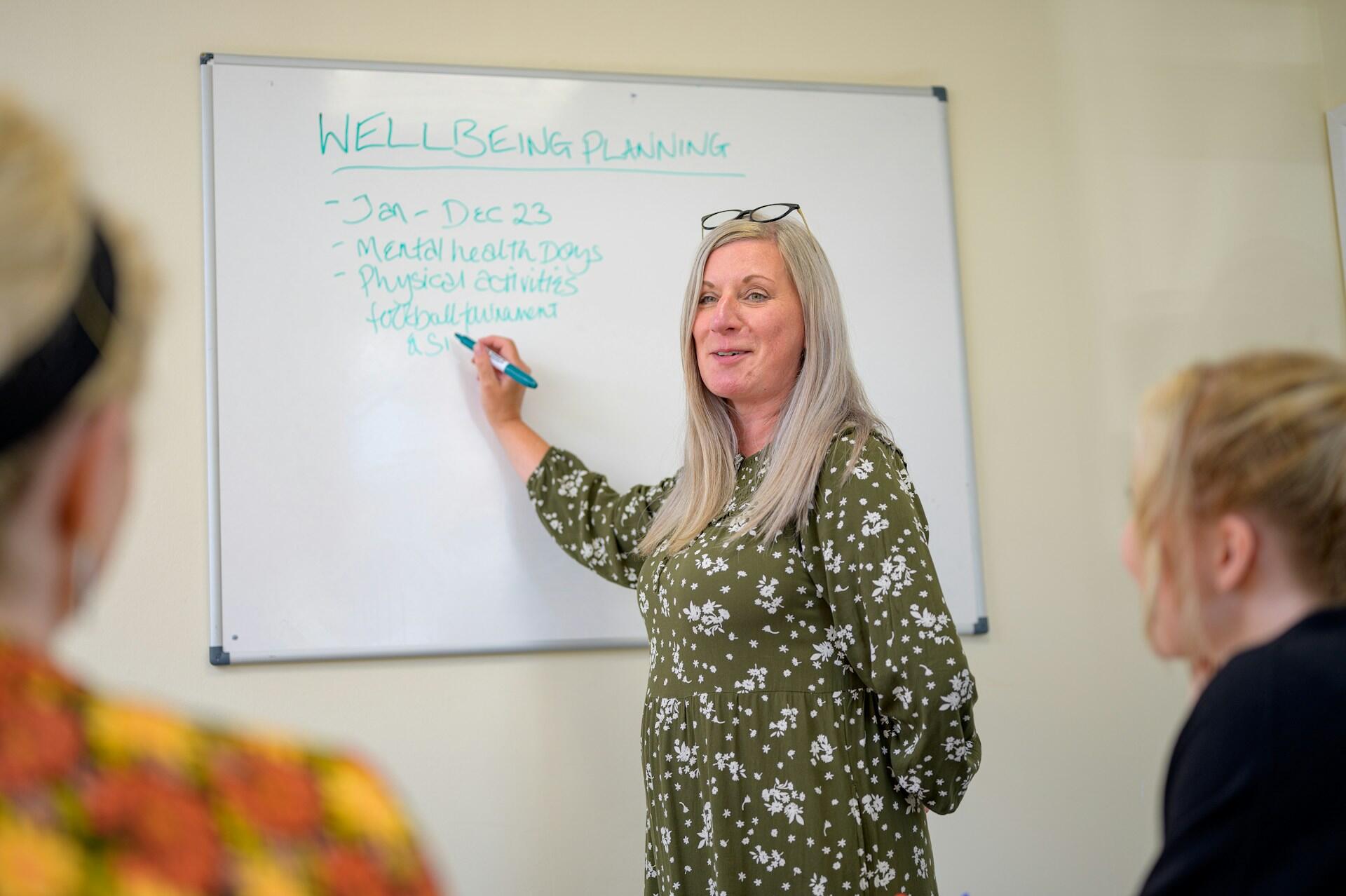 An older woman stands at a whiteboard pointing to writing on it while younger people sit facing the board. The mood is serious and encouraging.