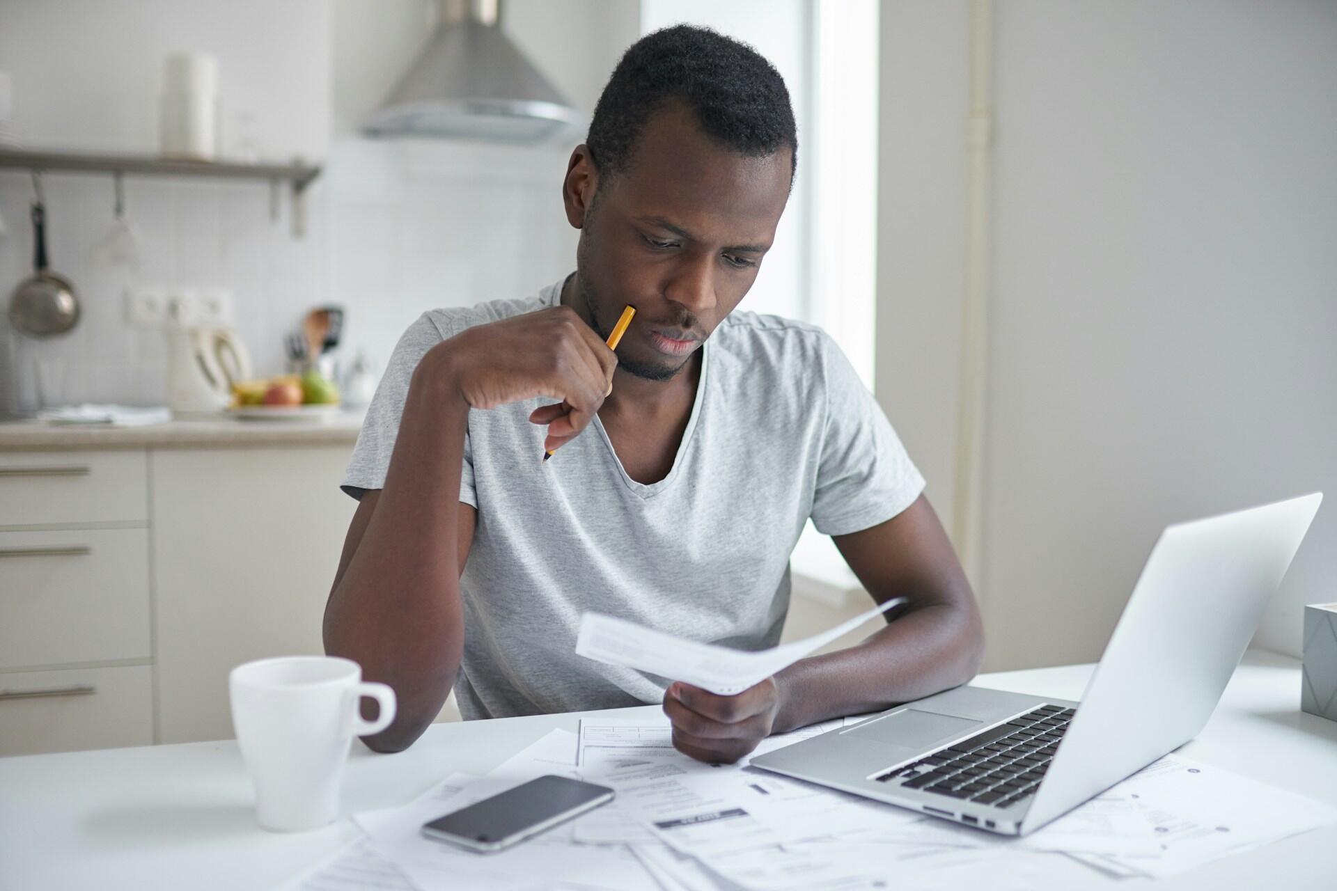 A man in a white shirt sits at a table, holding a piece of paper in his hand, with a pencil pointing at his face. He appears worried and thoughtful.