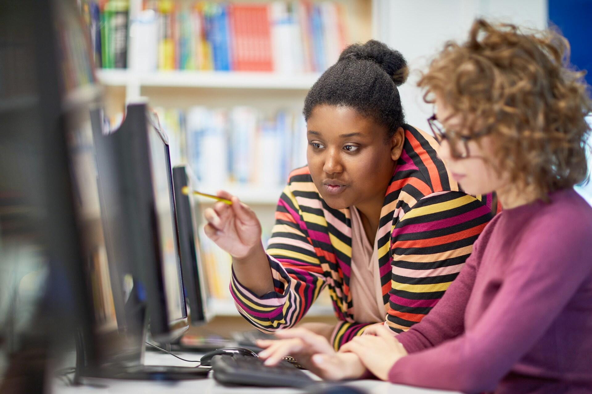 A woman wearing glasses, seated at a computer, looks down as another woman points to the screen. The mood is collaborative and helpful.