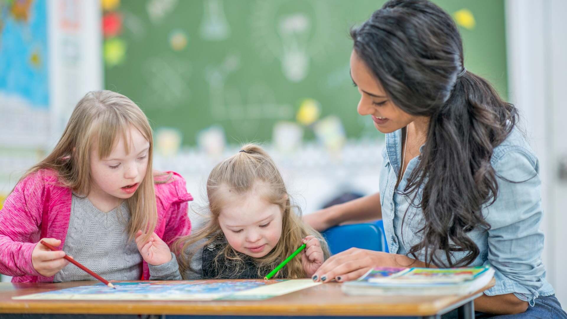 Δασκάλα βοηθά δύο μικρά κορίτσια να ζωγραφίσουν και να γράψουν σε τετράδιο μέσα σε σχολική αίθουσα.