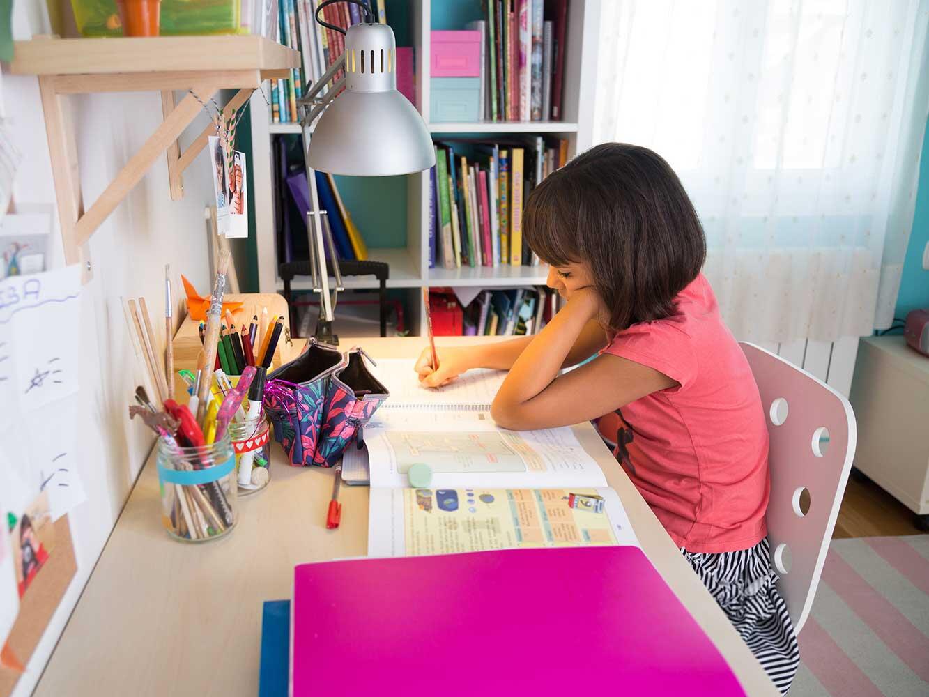 Κορίτσι κάθεται σε οργανωμένο παιδικό γραφείο και γράφει στο τετράδιό της, με σχολικά βιβλία και φωτιστικό γραφείου δίπλα της.