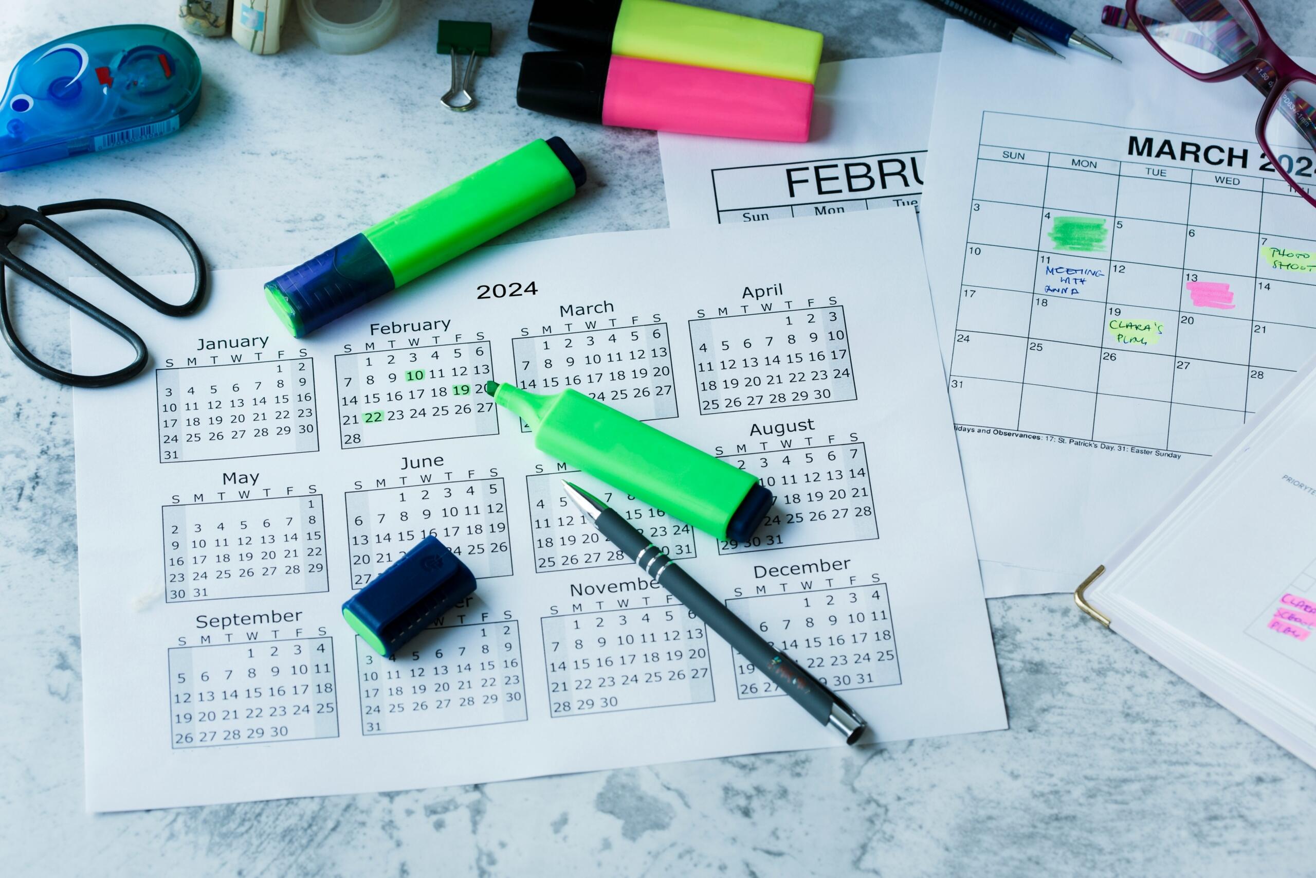 Εκτυπωμένο ετήσιο ημερολόγιο 2024 με σημειωμένες ημερομηνίες, μαρκαδόρους επισήμανσης και στυλό πάνω σε γραφείο, που δείχνει οργάνωση προγράμματος.