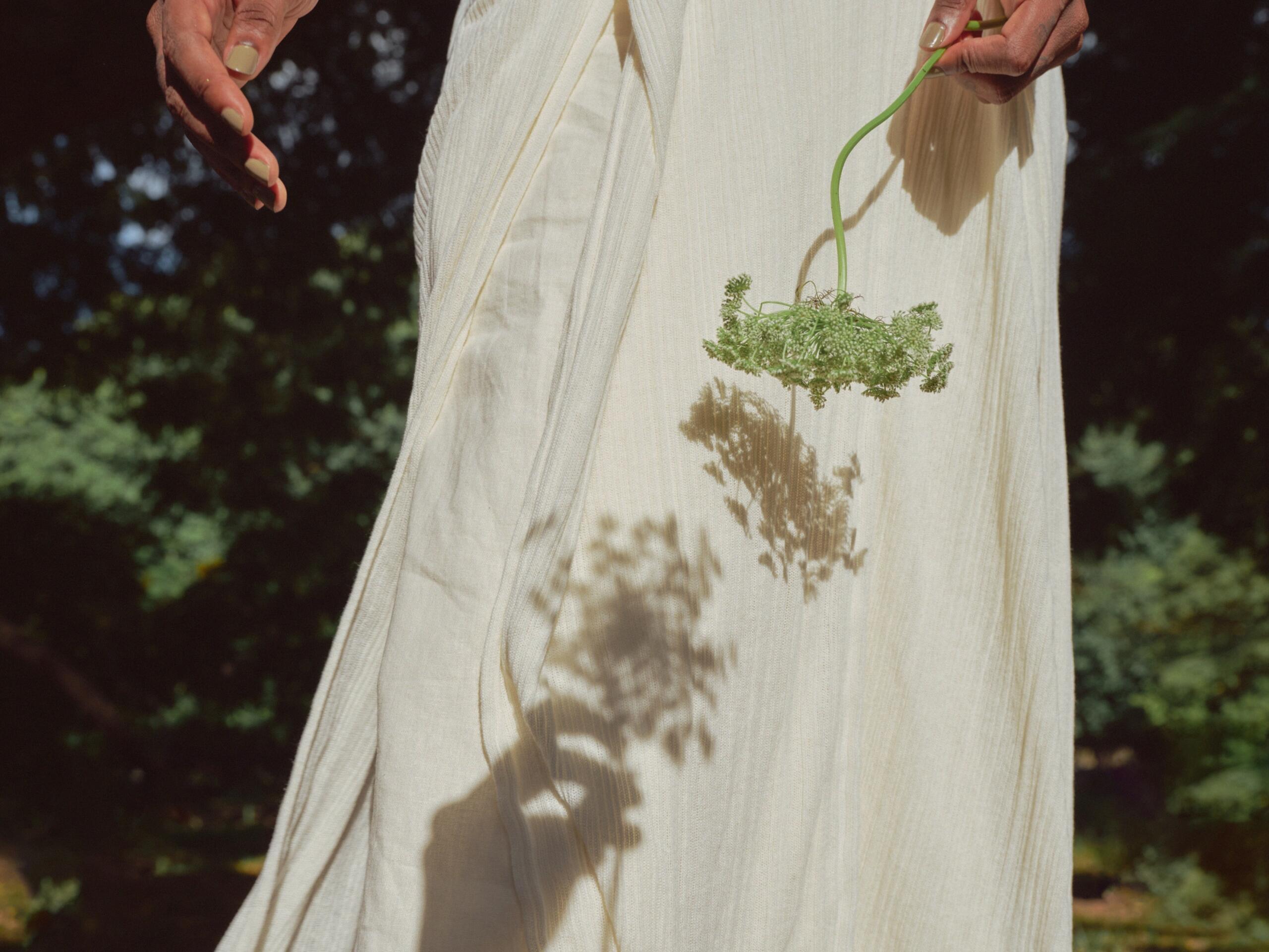 Άτομο με αρχαιοελληνικό ένδυμα κρατά φυτό σε εξωτερικό χώρο, με φυσικό φως και σκιά.