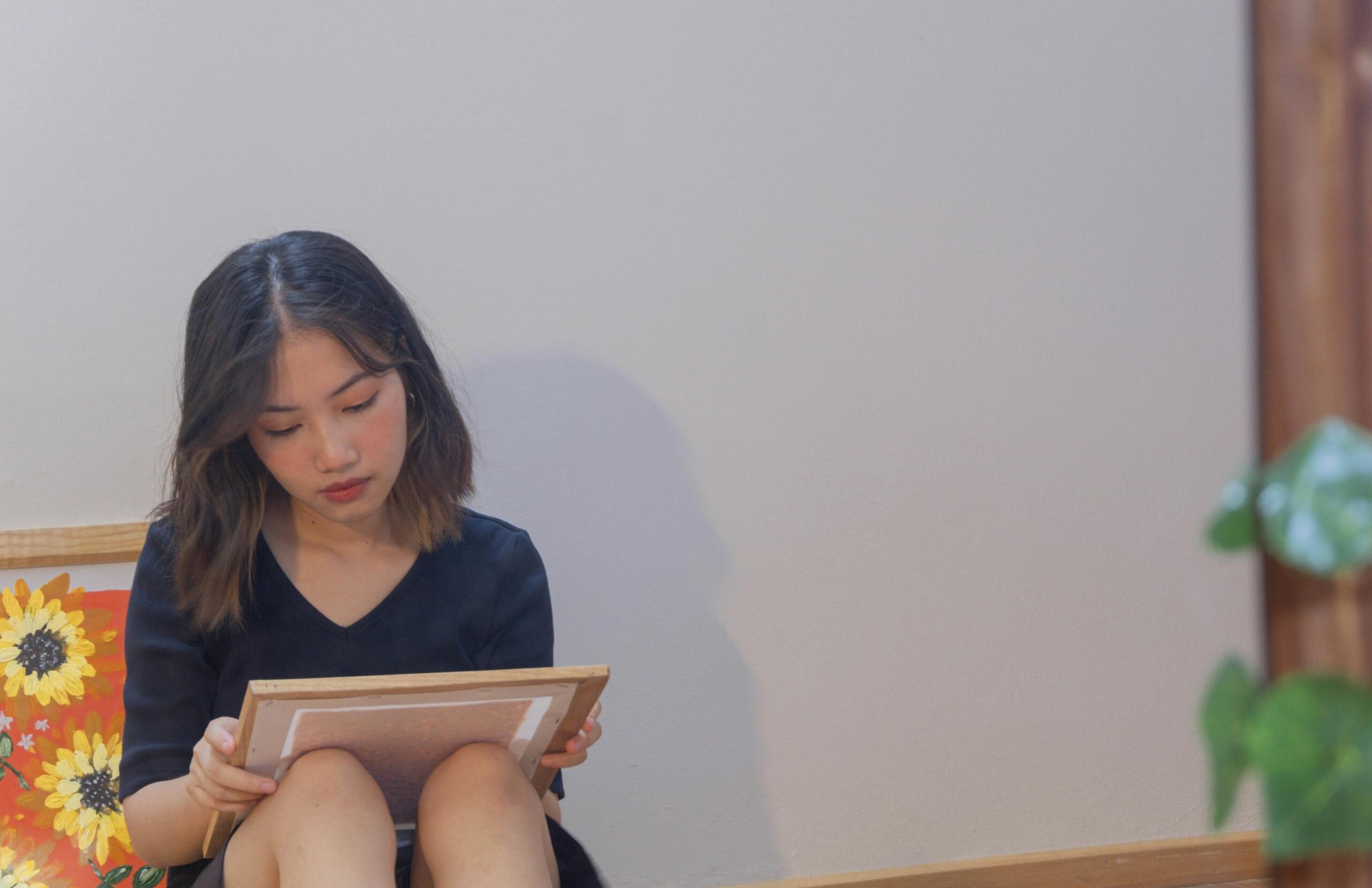 Νεαρή γυναίκα κάθεται σε πάγκο και κοιτάζει ένα πλαίσιο ή πίνακα που κρατά στα χέρια της.