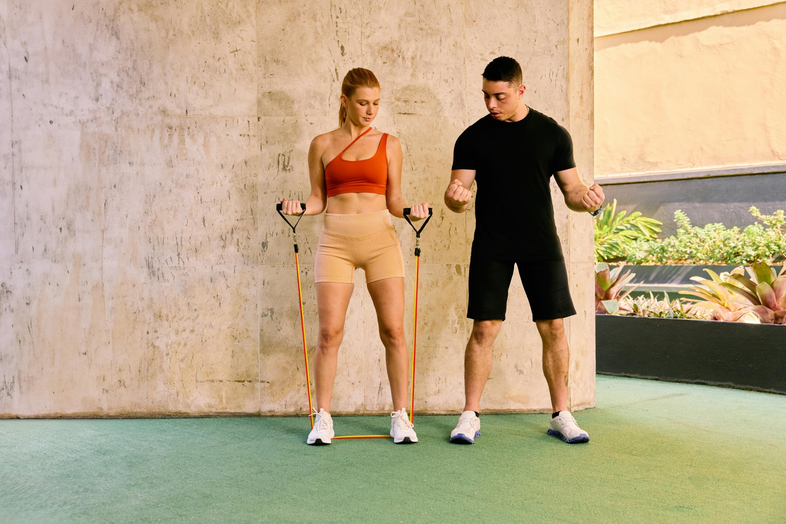Προπονητής δείχνει σε γυναίκα πώς να εκτελέσει άσκηση με λάστιχα αντίστασης.