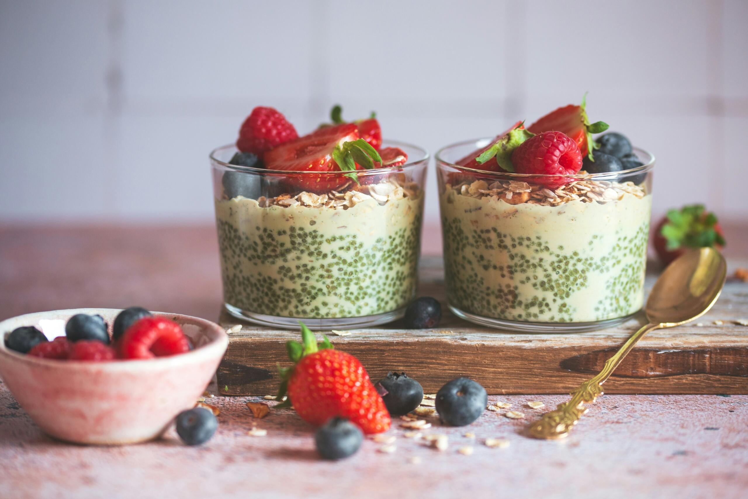 Δύο ποτήρια με vegan chia pudding, φυτικό γιαούρτι και επικάλυψη από φρέσκες φράουλες, σμέουρα, μύρτιλα και δημητριακά πάνω σε ξύλινη επιφάνεια.