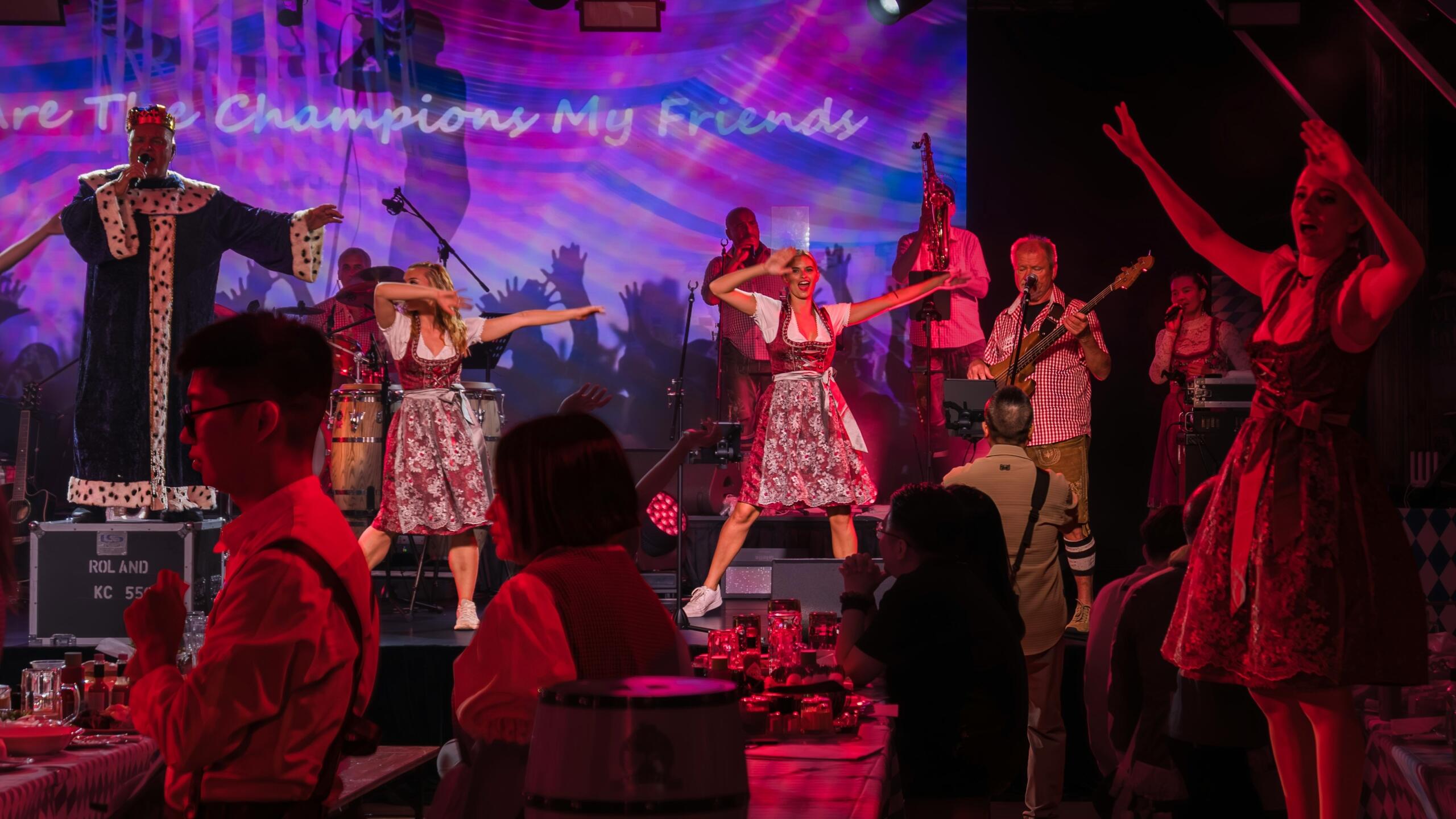 Ζωντανή σκηνή συναυλίας με χορεύτριες σε παραδοσιακές βαυαρικές φορεσιές και μουσικούς που παίζουν μπροστά σε κοινό μέσα σε φωτισμένο χώρο εκδήλωσης.