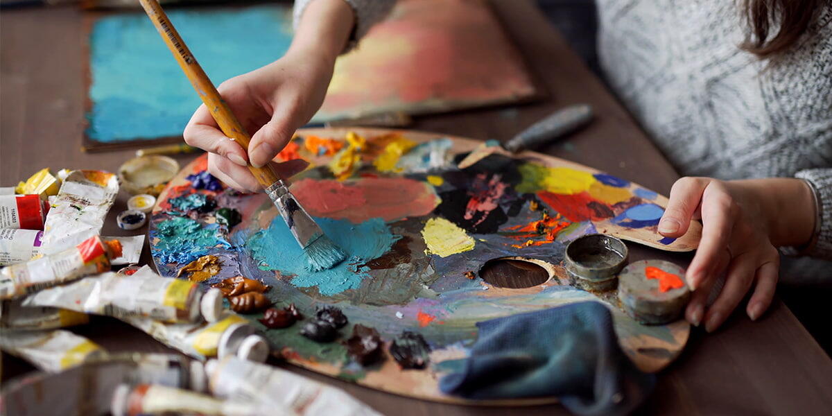 Κοντινό πλάνο χεριών που αναμειγνύουν πολύχρωμα χρώματα σε μια ξύλινη παλέτα, που περιβάλλεται από σωλήνες και πινέλα.