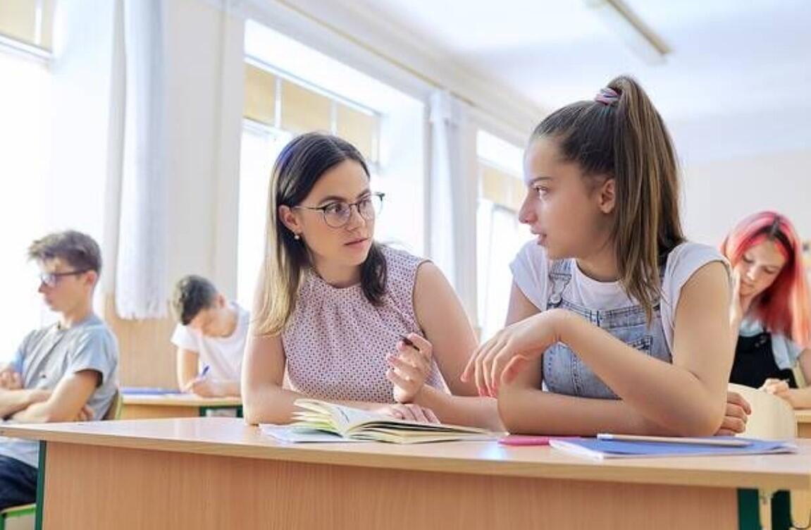 Καθηγήτρια βοηθά μαθήτρια μέσα σε σχολική τάξη, ενώ άλλοι μαθητές γράφουν στα θρανία τους.