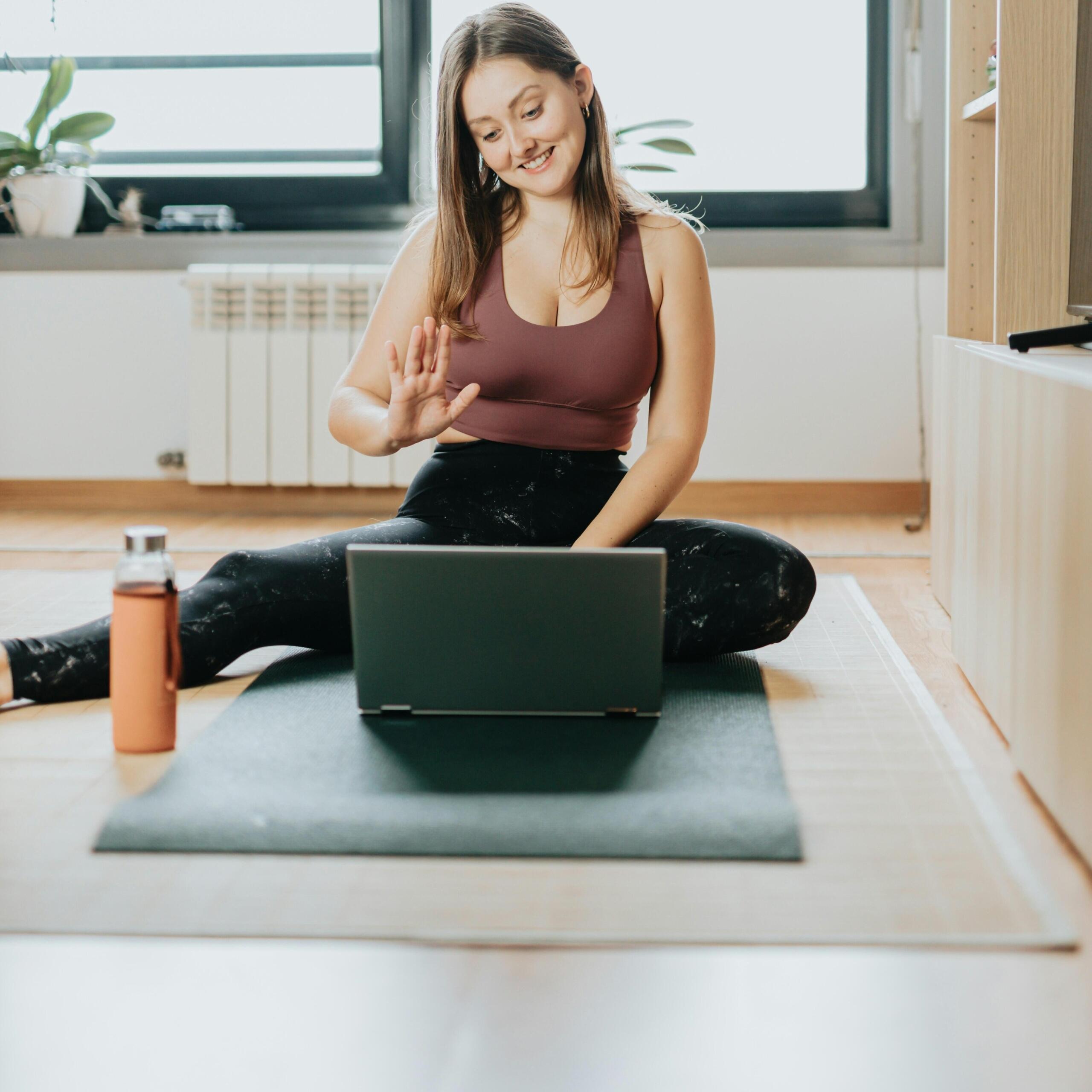 Γυναίκα κάθεται σε στρώμα γυμναστικής και παρακολουθεί διαδικτυακό μάθημα μέσω laptop στο σπίτι.