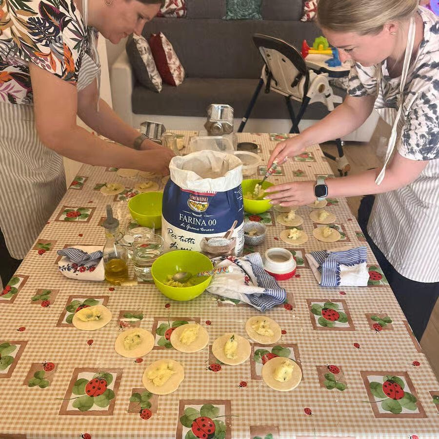 Δύο γυναίκες προετοιμάζουν ζύμη σε κουζίνα, γεμίζοντας μικρά φύλλα με γέμιση πάνω σε τραπέζι με υλικά μαγειρικής.