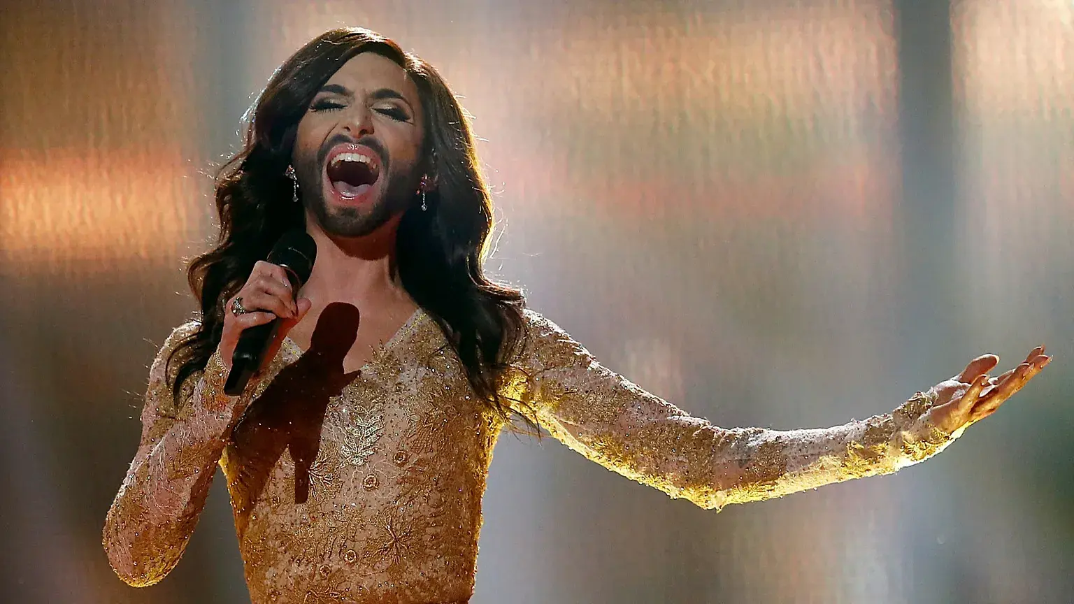 Η Conchita Wurst στη σκηνή της Eurovision με χρυσό φόρεμα, κρατώντας μικρόφωνο και τραγουδώντας με έντονη έκφραση κάτω από ζεστό φωτισμό.