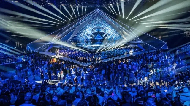 Μεγάλη σκηνή της Eurovision σε σχήμα πυραμίδας με εντυπωσιακά φώτα και πλήθος θεατών γύρω από τη σκηνή κατά τη διάρκεια ζωντανής εμφάνισης.