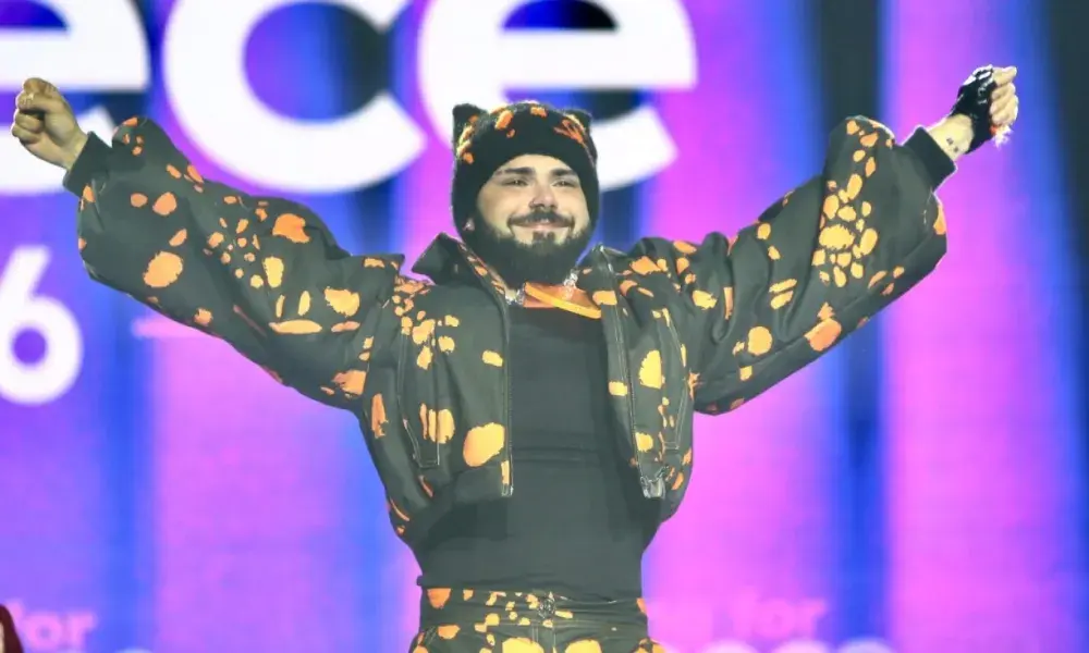 Ο Ακύλας στη σκηνή με μαύρο και πορτοκαλί outfit, σηκωμένα χέρια και χαμόγελο μπροστά σε φωτισμένο φόντο.